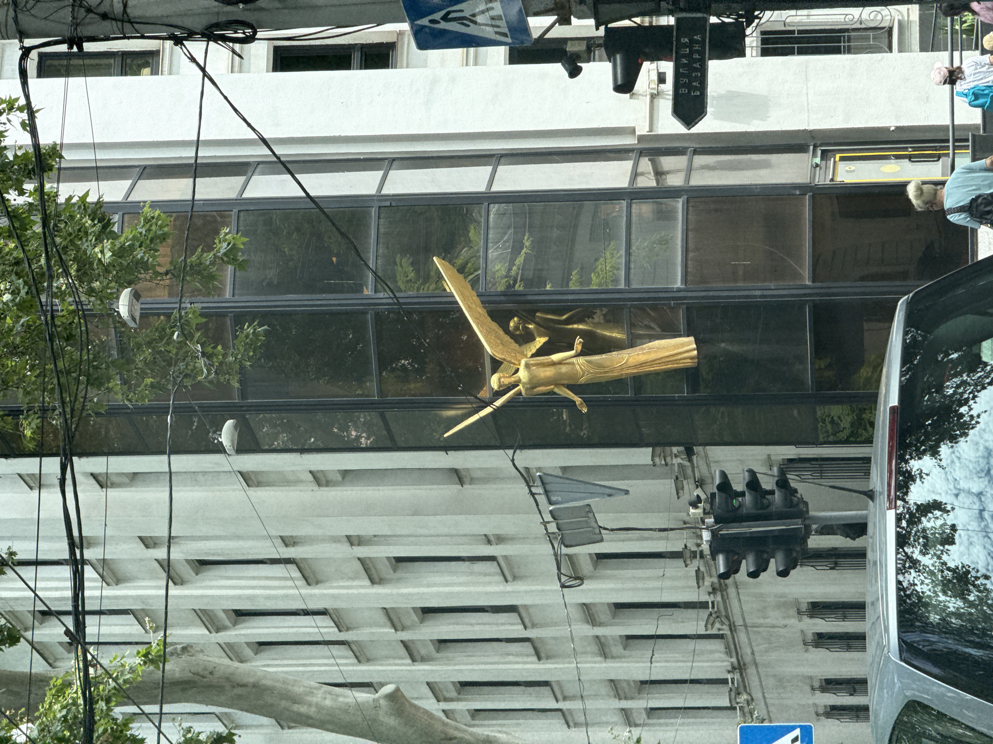 This media file captures a street view in Odesa, Ukraine. The primary subjects are a building with large glass windows, a golden statue of an angel with wings spread, and a traffic light. In the background, a portion of a silver vehicle is visible. Several people are present, including a woman with her hair in a bun wearing a light blue top and a black bag, and another person in the background wearing a white shirt and blue bag. A street sign with Cyrillic text reading "ВУЛИЦЯ БАЗАРНА" (Vulitsya Bazarna, meaning Bazarna Street) is also visible. The scene appears to be taking place during the daytime, under what seems to be overcast or slightly hazy weather, judging by the diffused light and reflections. The image is taken from a low angle, looking upwards at the building and the statue. The overall impression is of an urban environment with architectural elements and public infrastructure.