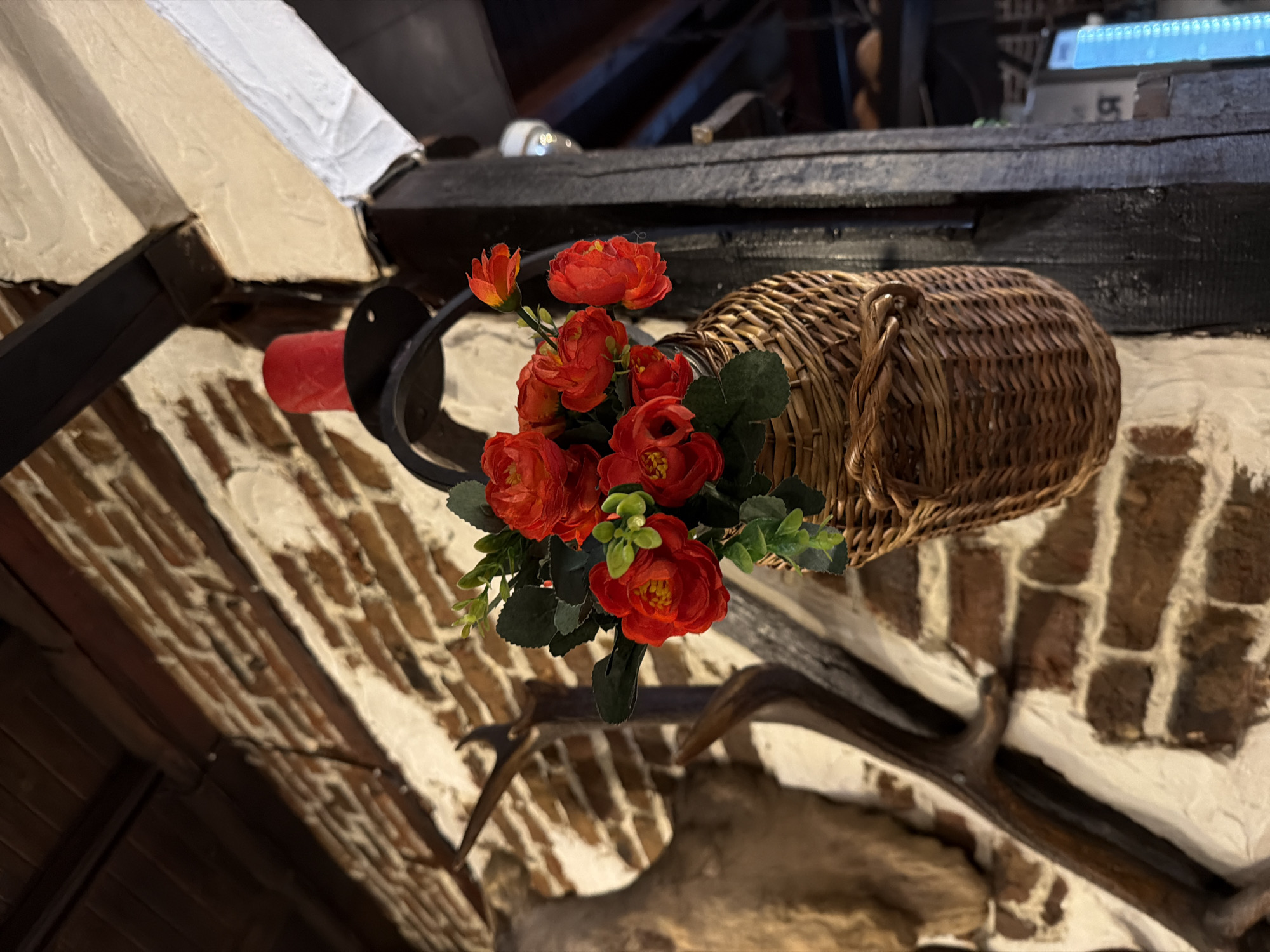 The image presents an interior scene, located in the city of Varna, Bulgaria. No individuals are present, and no actions or interactions are depicted.
Central to the composition, a woven wicker basket filled with artificial red flowers and green foliage hangs from a black metal bracket. A red cylindrical candle is seated within a holder atop this bracket. The bracket assembly is affixed to a dark, rough-hewn vertical wooden beam.
To the left of this central arrangement, a wall constructed from red bricks with white mortar is visible, partially covered by a light-colored plaster or whitewash. Mounted on this wall, the antlers and a portion of a taxidermy ungulate head are discernible.
In the upper left corner, dark wooden planks form a ceiling or overhead structure. To the right of the central wooden beam, another dark wooden beam runs vertically. Further to the right and slightly blurred in the background, a white cylindrical fixture is mounted on an overhead beam, and a vertically oriented, illuminated blue display or screen is partially visible. The overall environment suggests a rustic or traditionally decorated establishment.