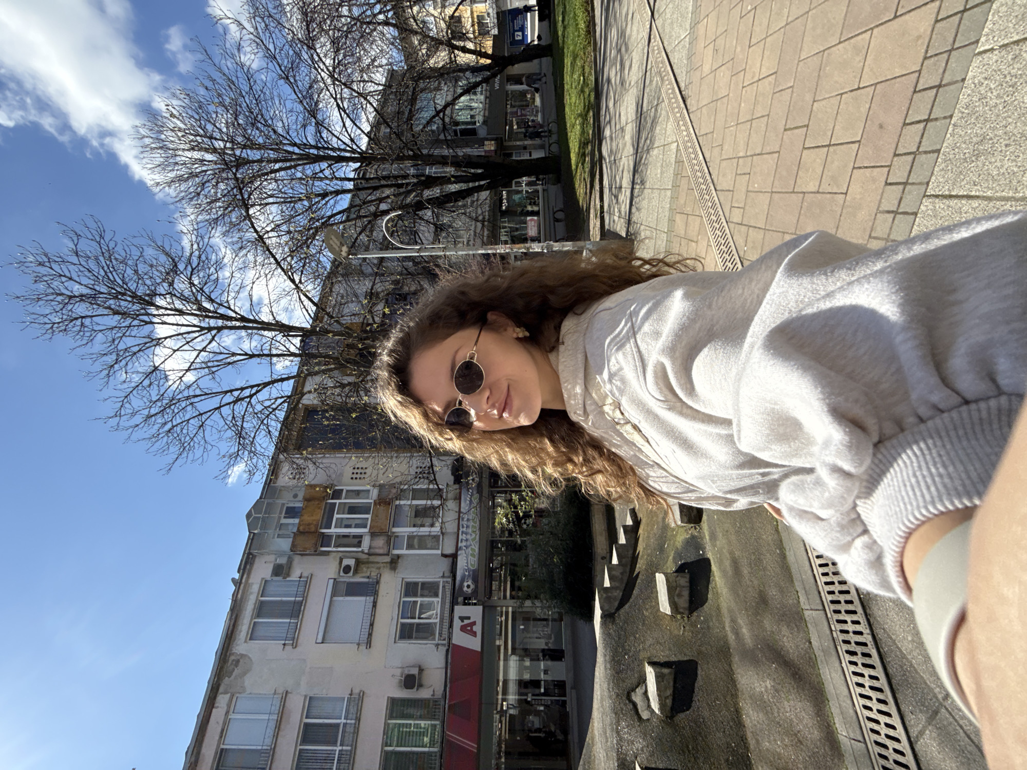 A medium outdoor shot shows a young, fair-skinned person in a light grey sweater and round sunglasses taking a selfie. The person is standing on a grey concrete walkway in Razgrad, Bulgaria. Behind the person is a multi-story building with multiple windows and the logo of the telecom "A1". Bare trees are above and to the left of the building. The sky is blue with some white clouds.