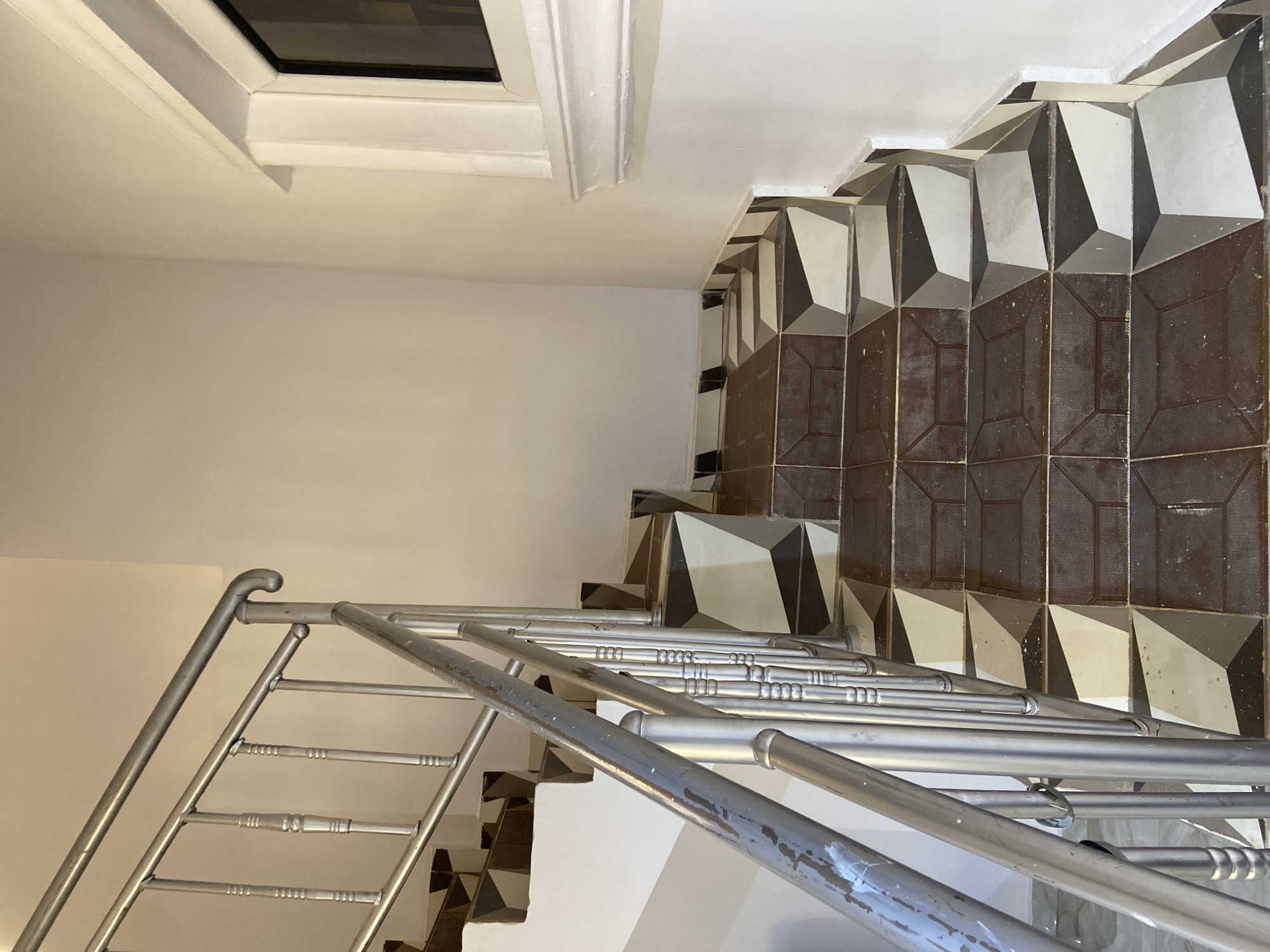 The image shows an indoor staircase with a silver metal railing. The steps are tiled with a pattern of dark brown and light gray geometric shapes, creating a 3D illusion. The walls are painted a light cream color. A white decorative molding runs along the wall near a window on the right side of the image. The scene appears to be a residential or commercial building in Gura Top, Nigeria. There are no people visible in the image.