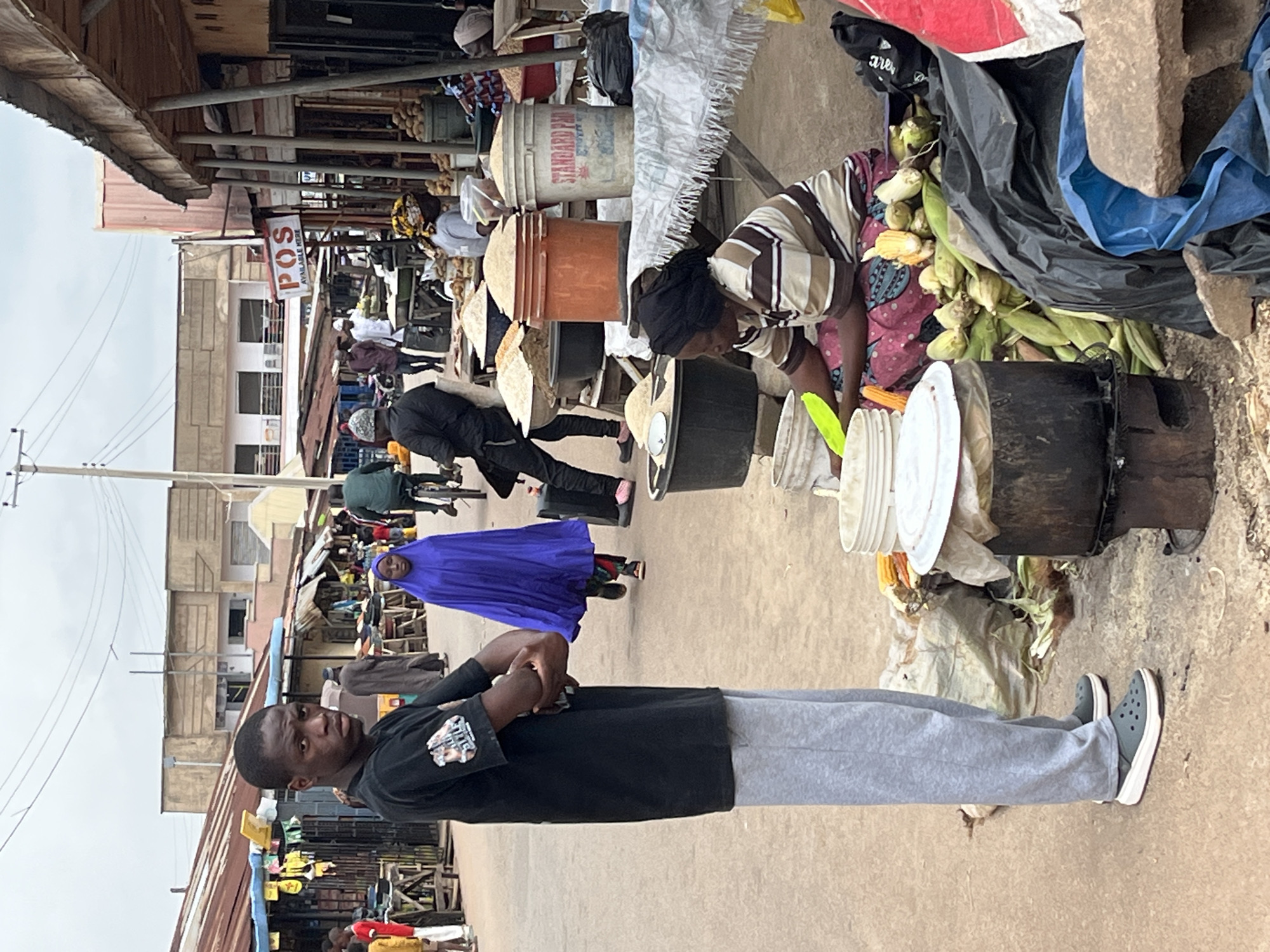 A street market scene unfolds in the city of Jos, Nigeria, under an overcast sky. A young Black man, standing prominently in the foreground and facing the camera with a direct gaze, wears a black t-shirt and grey sweatpants. His arms are crossed, and he sports dark green slip-on shoes. To his right, a woman in a vibrant purple hijab walks away from the camera, her figure partially obscured by the man. Further into the market, a woman kneels behind a table laden with corn cobs and other produce. She wears a striped shirt and a patterned skirt, with a black headscarf. Various buckets and containers filled with grains are visible on tables and the ground, indicating the market's nature as a place for agricultural goods. In the background, stalls with corrugated metal roofs line the street, and buildings with modern architecture rise above. The overall impression is a bustling, everyday scene in a Nigerian urban market.
