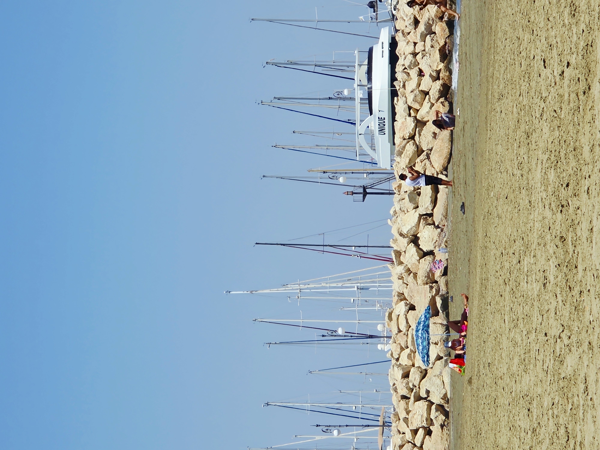 The image depicts a sunny day at a beach in Larnaca, Cyprus, with a clear blue sky. In the foreground, a sandy beach is visible, with a few people relaxing. Two individuals are lying under a blue and white umbrella, with bags and towels nearby. Further along the beach, a man stands and appears to be looking at his phone, while a woman crouches near the water's edge. Another woman is visible walking further down the beach.
Behind the beach is a breakwater constructed from large, light-colored rocks. Beyond the breakwater, a harbor is visible, filled with numerous sailboats and yachts. One prominent white yacht with "UNIQUE" written on its side is docked. The masts of the sailboats create a distinct line against the sky. The water in the harbor appears calm. The scene suggests a typical tourist or recreational atmosphere in a coastal city.