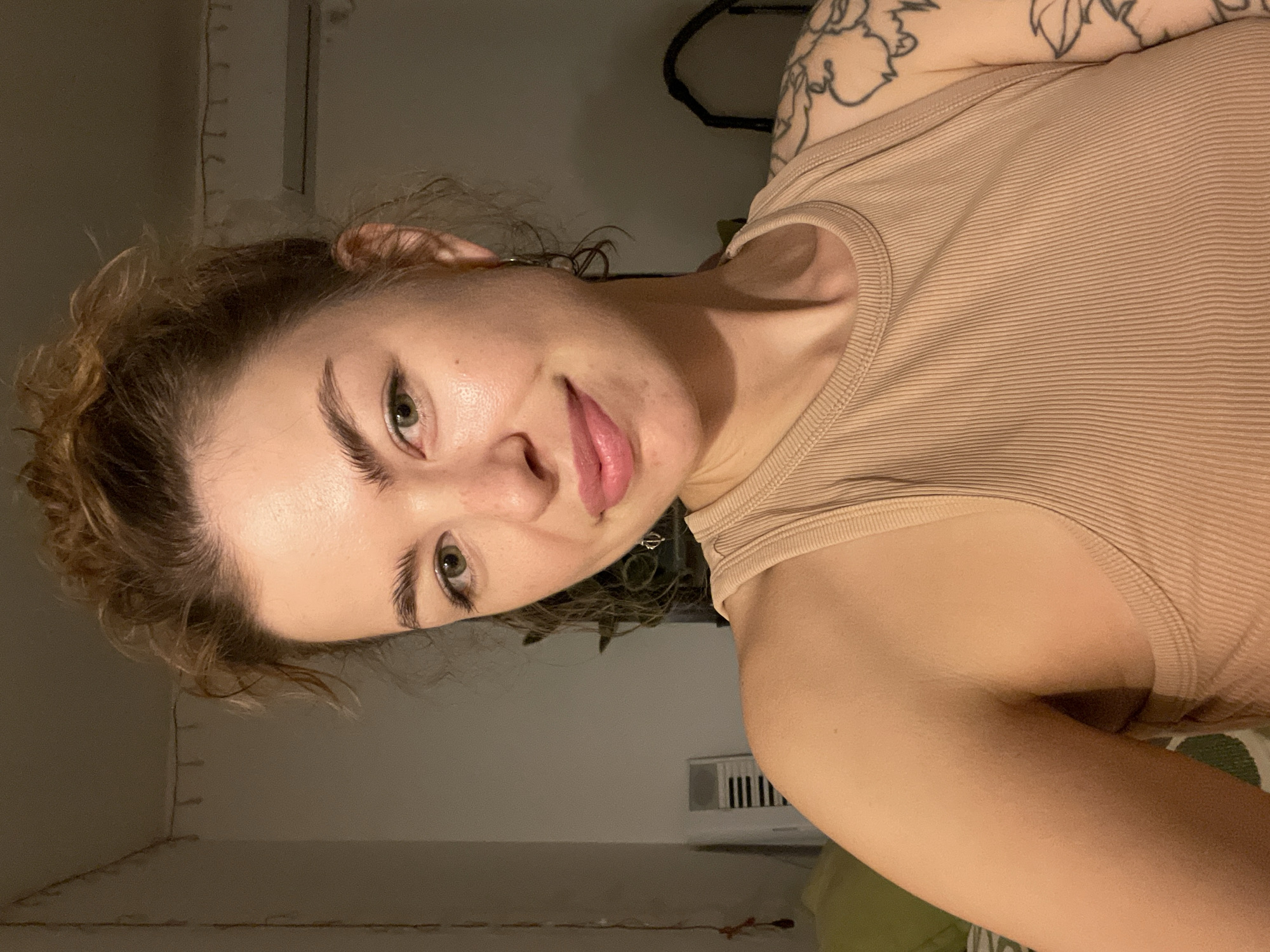 A selfie of a fair-skinned person with light brown, curly hair, green eyes, and pink lips, wearing a tan ribbed tank top. The person is facing the camera at a slight angle. A tattoo is visible on the upper right arm. The background includes a wall with string lights, a black cable, and a vent. The location is not specified.