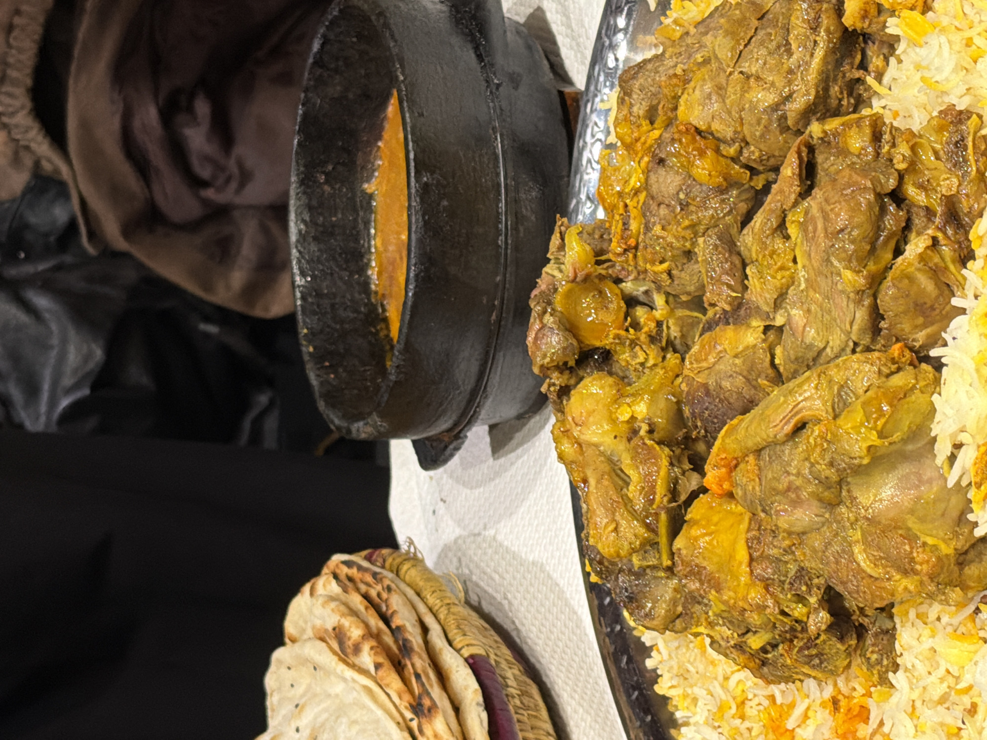 The image shows a close-up of a Middle Eastern dish, likely Majboos or Mandi, featuring a large serving of spiced rice topped with tender pieces of cooked lamb or mutton. The meat has a yellow-orange tint, probably due to saffron or turmeric used in the cooking. Next to the plate is a small, black pot containing a sauce, possibly a tomato-based or yogurt-based gravy. In the foreground, there is a basket filled with thin, flatbreads, likely Khubz or Markook. The scene is likely indoors, possibly in a restaurant or at a catered event.
Without additional information, it is impossible to determine if the image was taken in Paris, France. There is nothing in the image itself to indicate the location.