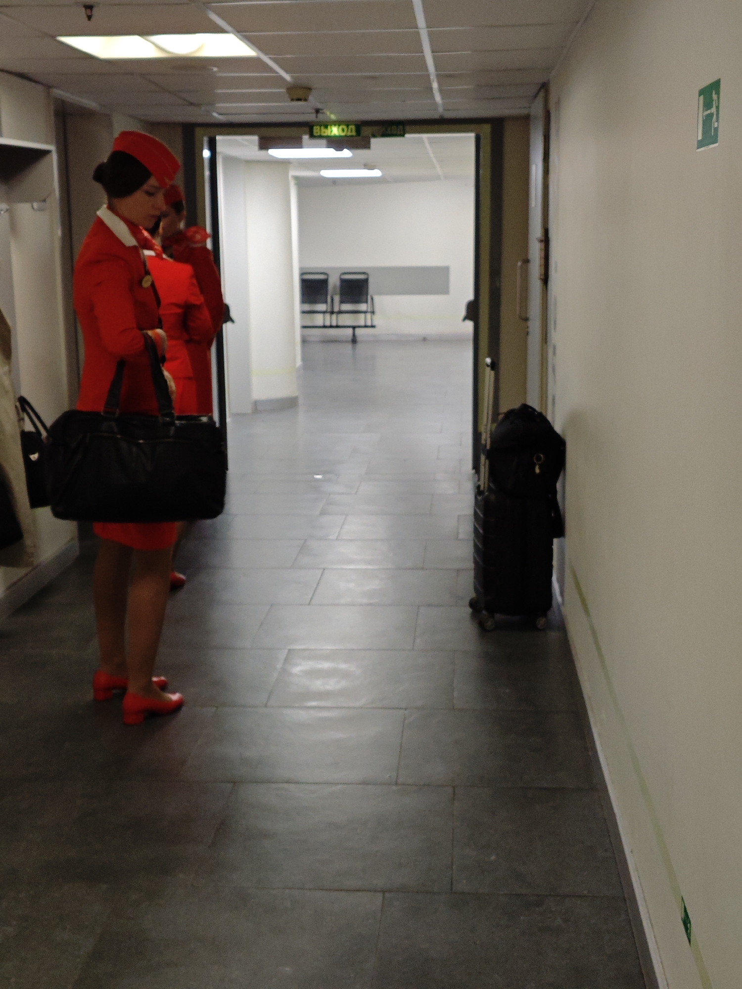 The image shows a dimly lit interior hallway with two individuals dressed as flight attendants in red uniforms. The flight attendant in the foreground is on the left, facing right, with a large black handbag in her left arm. The second flight attendant is to her right, also in uniform, partially obscured. In the background, a doorway leads to a brighter area with chairs and a sign that reads "ВЫХОД" (Exit). On the right side of the hallway, a black wheeled suitcase is propped against the wall. The floor is tiled, and the walls are painted a neutral color. A green sign with a white symbol is visible on the far right wall. The scene appears to be within an airport or similar transportation hub, possibly at Pulkovo Airport in Saint Petersburg, Russia, given the signage and overall environment.