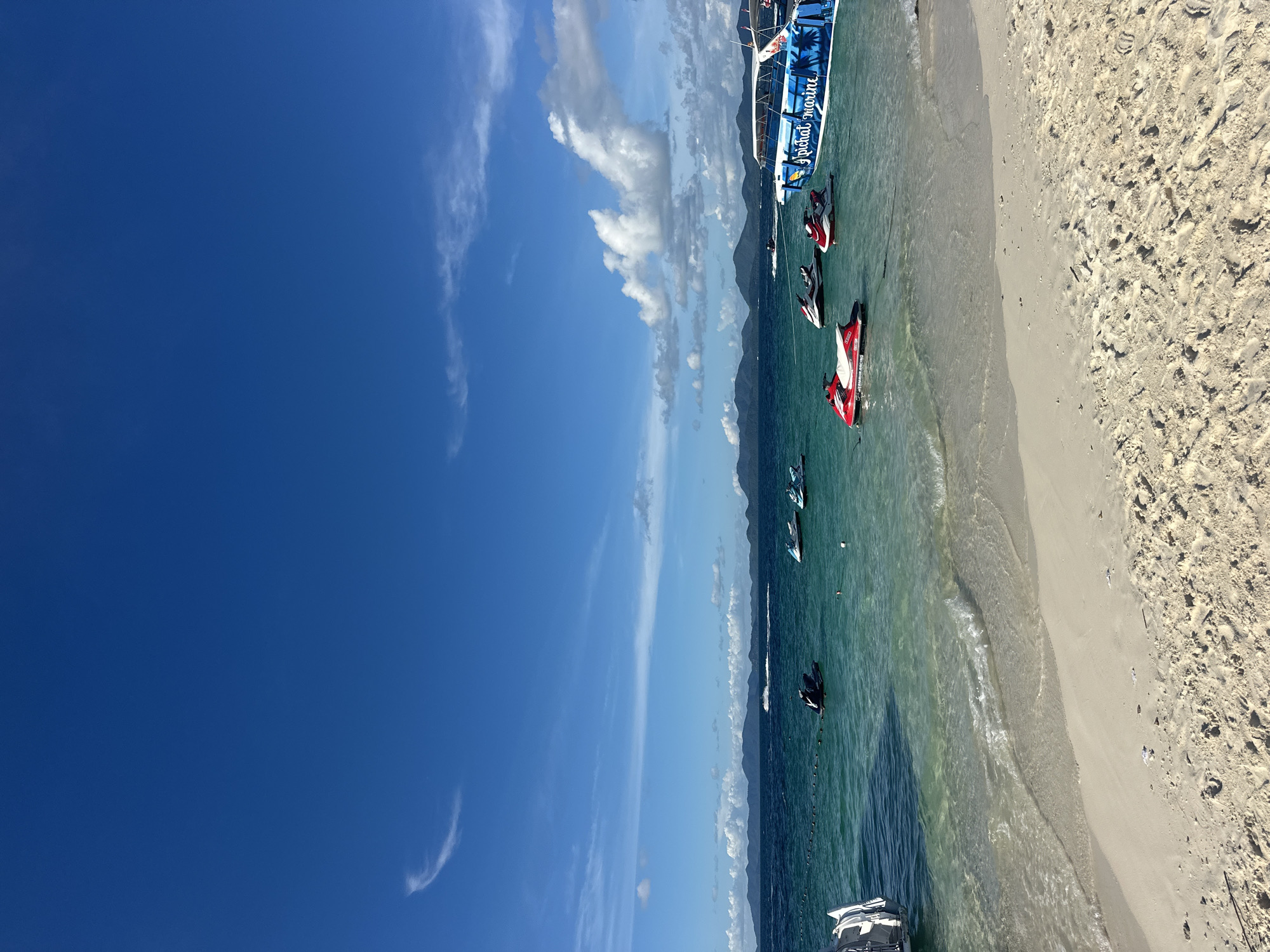This image captures a vibrant beach scene with a focus on watercraft and the serene ocean. In the foreground, a sandy beach transitions into the clear, turquoise water. Several jet skis are scattered across the water's surface, with some in motion and others stationary. The jet skis are predominantly red and white. In the background, a large boat with "Tropical Marine" written on its side is docked. The boat is adorned with palm tree decals, suggesting a tropical setting.
The water is calm, with gentle waves lapping at the shore. The sky above is a brilliant blue, dotted with scattered white clouds that are illuminated by the sun, indicating a bright, sunny day, likely midday given the lighting. In the distance, a mountain range can be seen, partially obscured by the clouds, adding depth to the landscape. The presence of jet skis and a tour boat implies that recreational activities are taking place, possibly a tourist excursion or rentals. The overall mood of the image is one of leisure and outdoor fun. The sandy beach, clear water, and sunny weather create a quintessential tropical vacation atmosphere. The visible text "Tropical Marine" on the boat clearly identifies the operator or service.