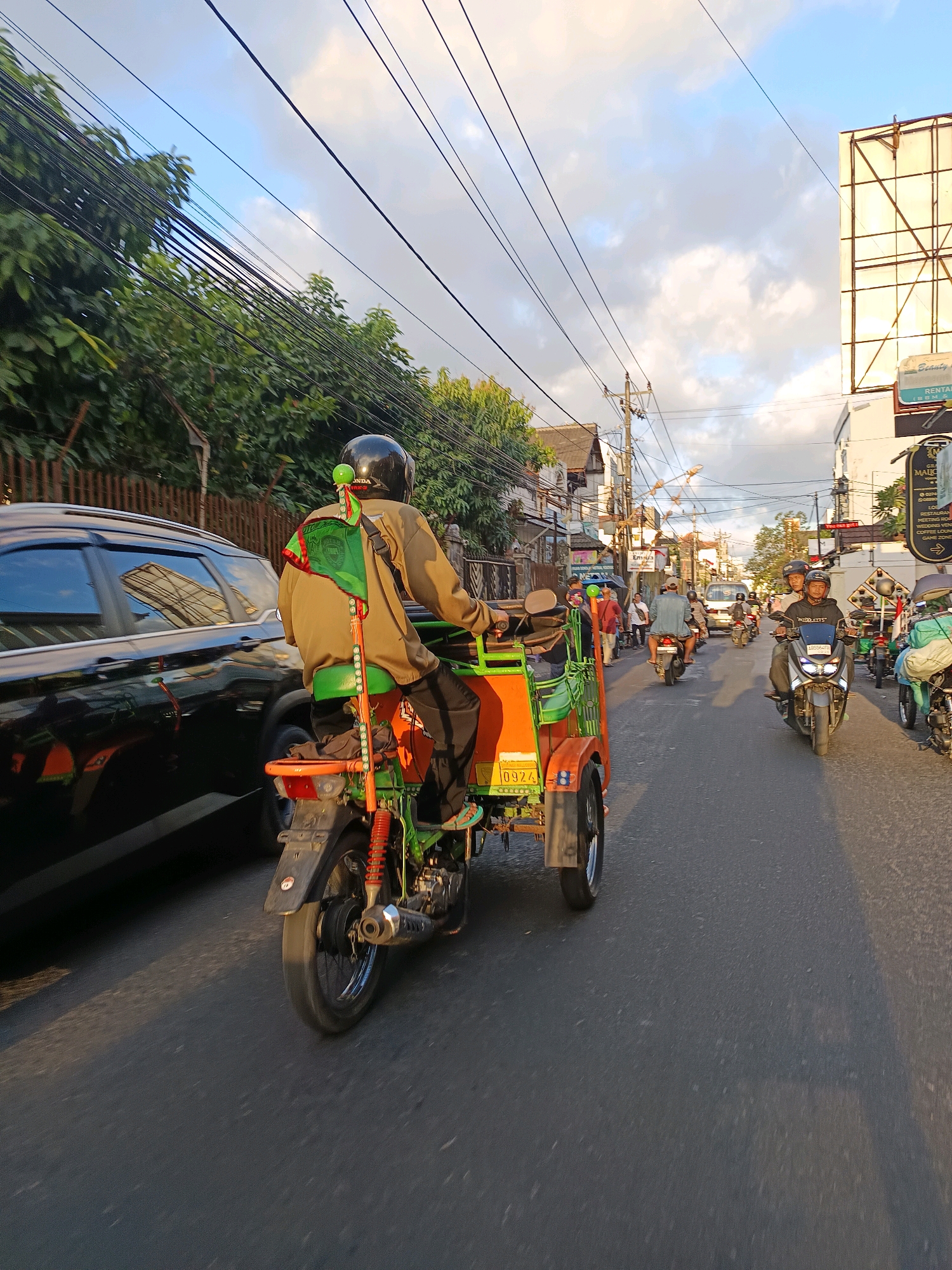 A man wearing a tan jacket and black helmet rides a colorful three-wheeled motorcycle, which has a green seat and orange trim, down a street in Yogyakarta, Indonesia. The motorcycle has a green flag attached to a pole at its rear. The man is riding in the right lane of a two-way street. A black SUV is to his left, and other motorcycles and mopeds are traveling in both directions on the street. The sky is a mix of blue and clouds, and overhead power lines crisscross the scene. Buildings, trees, and a large billboard are visible along the sides of the street.