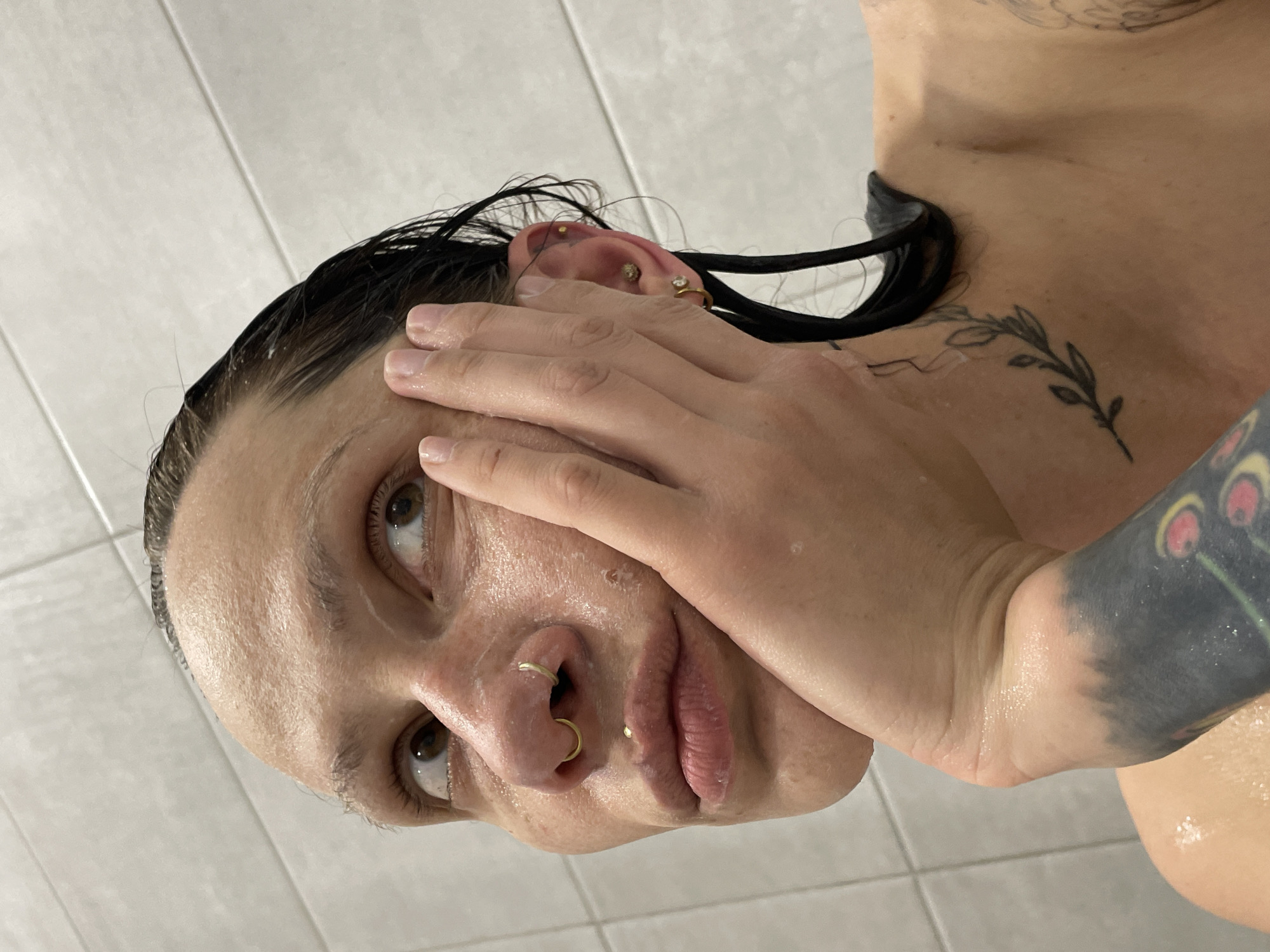A person is pictured close-up in what appears to be a shower. The person has short dark hair and is holding their hand up to their face. They have multiple piercings in their ear and nose. A tattoo is visible on their chest and arm. The background is a tiled wall. There is no indication of the location being Izhevsk, Russia.