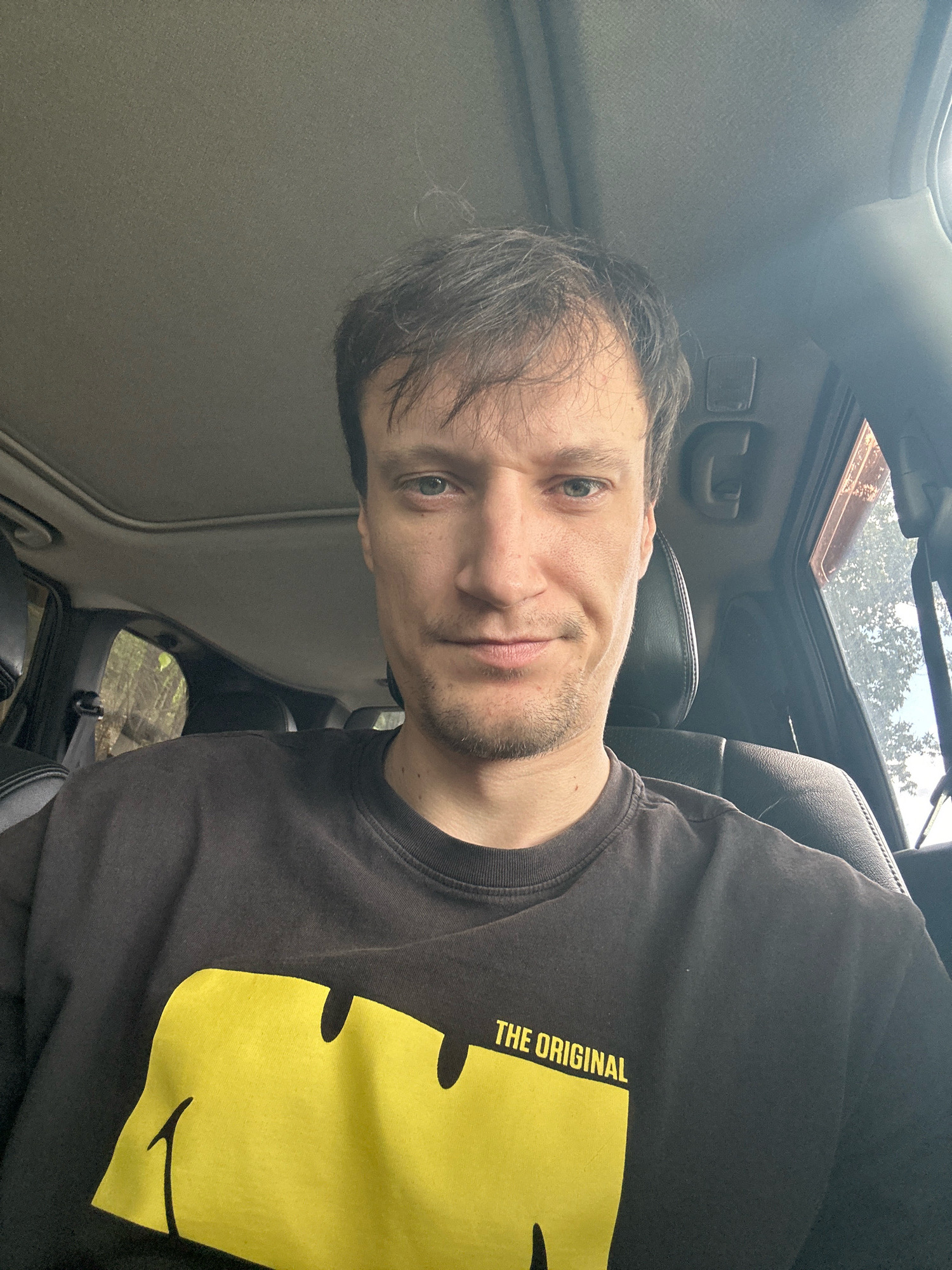 A man is seated in the driver's seat of a vehicle, likely in Makiv, Ukraine, based on context, with a selfie perspective. He has short, brown hair, light-colored eyes, and a light beard. He is wearing a dark-colored t-shirt with a large yellow graphic design and the words "THE ORIGINAL" printed on it. The interior of the vehicle is visible, including the car's sunroof, leather seats, and a portion of the passenger side window. Outside the window, trees and foliage are visible, suggesting a rural or natural setting. The lighting indicates it is daytime.