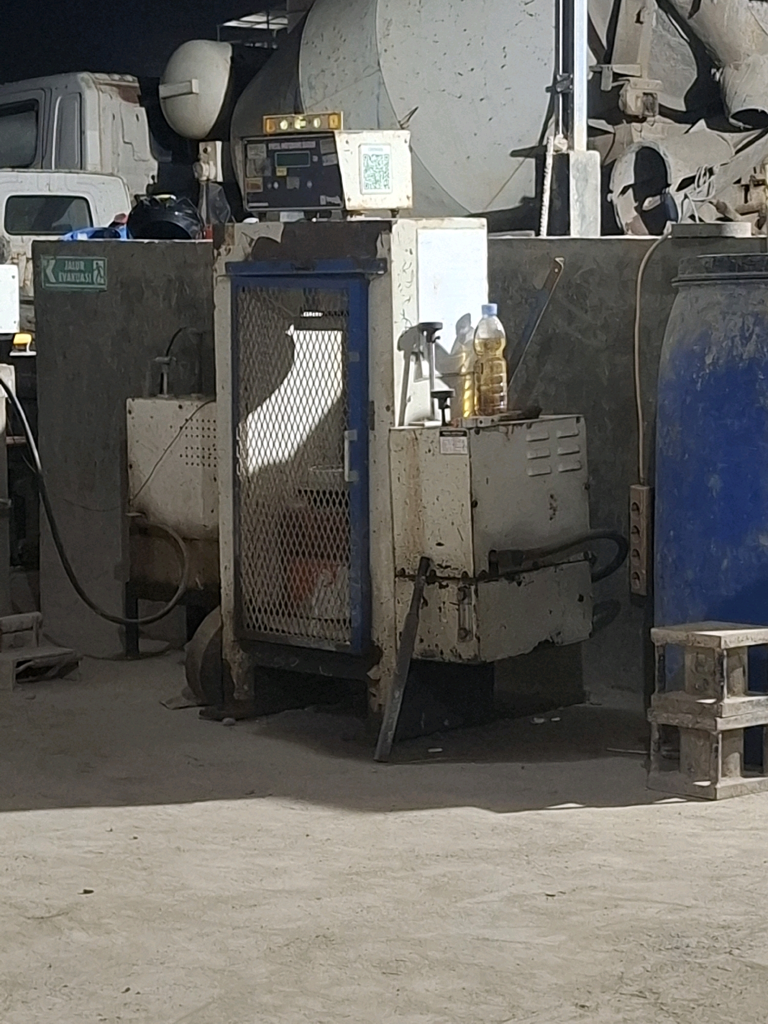 The image depicts an industrial setting, possibly a workshop or a factory, in Kutasirna, Indonesia. In the foreground, there is a large blue barrel on the right, and to its left, a white machine with a caged front enclosure. Inside the enclosure, a partially obscured figure is visible, possibly a person or a large object. Two bottles of yellow liquid are placed on top of the white machine.
Behind the white machine, there is a concrete structure with a green sign that reads "JALUR EVAKUASI" (Evacuation Route). Further back, several large industrial machines and parts of vehicles, including the cabin of a white truck, are visible. The environment appears dusty and cluttered, consistent with an operational industrial area. The lighting suggests it might be late afternoon or early evening. The ground is paved with concrete.