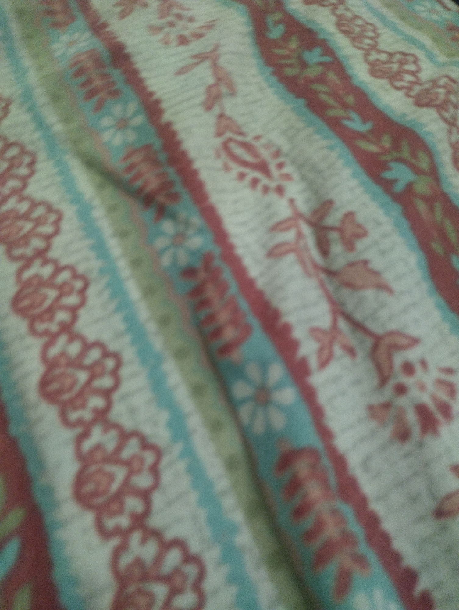 The image shows a close-up, slightly out-of-focus view of a fabric with a patterned design. The pattern consists of vertical stripes of varying colors and textures. One prominent stripe is a deep red with a zig-zag edge, adorned with light blue leaf-like shapes. Adjacent to this are stripes of light blue with white floral motifs and a cream-colored stripe with a repeating reddish-pink floral or paisley design. The fabric appears to be soft, possibly a bedsheet or a similar textile. There are no discernible people, actions, or environmental features of a city. The location is Unknown, Unknown.