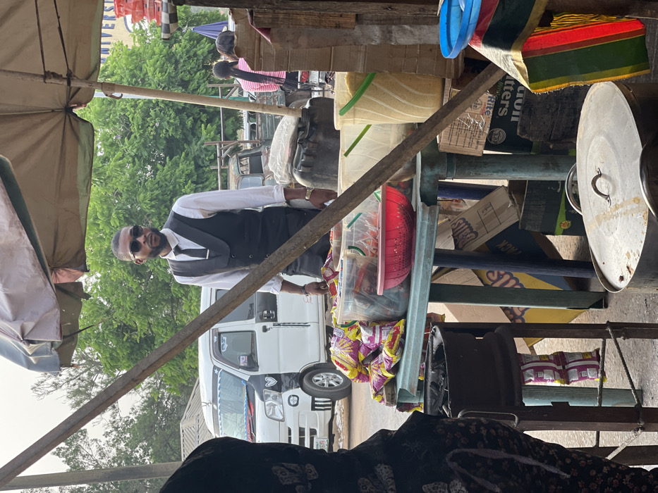 The image captures an outdoor market or street vending scene during what appears to be bright daylight. The perspective is from a low angle, looking towards a vendor and his stall.
In the right half of the image, a man stands attending to his goods. He is dressed in a white long-sleeved shirt, a dark vest, and a dark tie, suggesting a somewhat formal appearance for a street vendor. He has a beard and is wearing dark sunglasses, obscuring his eyes. His right hand is visible, resting on or near items on his stall.
The stall itself is a rustic setup, constructed from metal poles and wooden planks. It holds various items for sale. On the left side of the stall (from the viewer's perspective), there are transparent plastic containers; one appears to be filled with light-colored granular material, possibly rice or other grains, while another has a red lid. Below these, multiple plastic bags filled with snacks or chips, featuring yellow and purple packaging, are displayed. On the right side of the stall, a large metal pot with a lid is visible. Above it, a blue plastic basin and a piece of striped fabric (with green, yellow, and red colors) are placed. Several cardboard boxes are stacked behind the main display, with partial text visible, including "Lion Butters," "RITE," and "Dais."
In the background, behind the vendor and his stall, green trees indicate an outdoor setting. Several vehicles are parked or moving; a white van or mini-bus is prominently visible. Other people are faintly discernible in the upper left background, including one person with long, braided hair.
In the immediate foreground, at the bottom of the image, the back of a person wearing a dark, patterned garment is visible, suggesting the photograph was taken from behind another individual who might be seated or observing. The overall atmosphere suggests a typical bustling market environment.