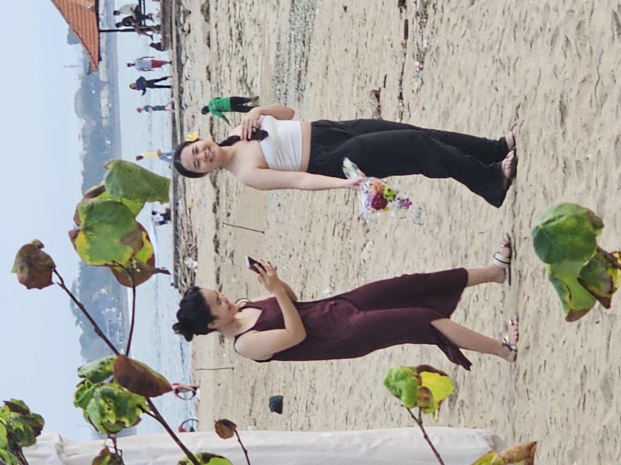 In Denpasar, Indonesia, two young women walk on a sandy beach near the ocean. The woman on the left, wearing a dark maroon slip dress, looks down at her smartphone, holding it in her right hand. She wears flat sandals. The woman on the right, in a white strapless top and black pants, smiles and holds a bouquet of colorful flowers in her left hand. A yellow flower is tucked behind her ear. In the background, the ocean stretches to the horizon, with a few buildings and palm trees visible on the distant shore. Several other people are scattered on the beach, some sitting, others walking. A small red-roofed structure is on the right side of the frame. In the foreground, out-of-focus green leaves and branches partially obscure the view, suggesting the photo was taken from behind foliage. The scene appears to be a casual, outdoor setting on a cloudy day.