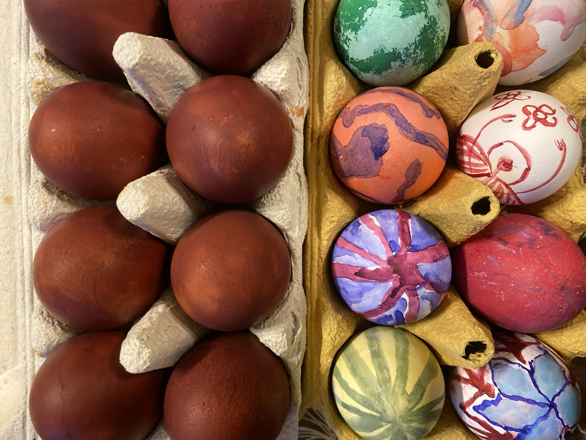 The image shows two egg cartons. The left carton contains six red-brown eggs, while the right carton contains eight eggs decorated with different colors and patterns including green, orange, purple, white, red, and blue. The egg carton is placed in an indoor setting. The location of the image is unknown. There is no evidence in the image that the eggs were painted in Moscow, Russia.