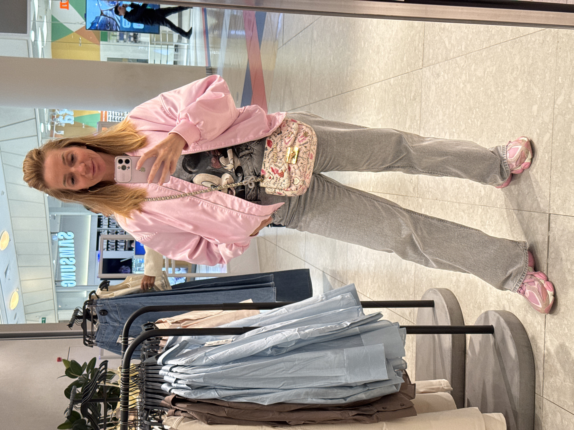 This media file features a mirror selfie of a woman taken inside what appears to be a shopping mall or large retail store in Saint Petersburg, Russia.
The primary subject is a woman with shoulder-length blonde hair and gold hoop earrings. She is wearing a light pink bomber jacket over a gray t-shirt with a Mickey Mouse graphic, light gray loose-fitting jeans, and distinct chunky pink and white sneakers. She carries a small, light pink patterned shoulder bag with a gold chain strap and a gold clasp. The woman is holding a light purple smartphone, likely an iPhone, with a clear case, reflecting her image in a large mirror. She has a pleasant, slightly smiling expression, suggesting a casual and relaxed mood.
The setting is an indoor commercial space with a light-colored tiled floor and large white pillars. In the foreground and background, there are clothing racks displaying various garments, including folded light blue and brown shirts, and hanging denim pants. The activity is the woman taking a mirror selfie, likely documenting her outfit or outing.
Notable details include reflections in the mirror: a reversed sign that reads "SNASMUS" (SAMSUNG), indicating an electronics store or section nearby, and a blurred reflection of another person walking past a large screen. The overall atmosphere is that of a modern retail environment during what appears to be daytime, with artificial lighting. The woman's outfit showcases a casual and trendy style.
