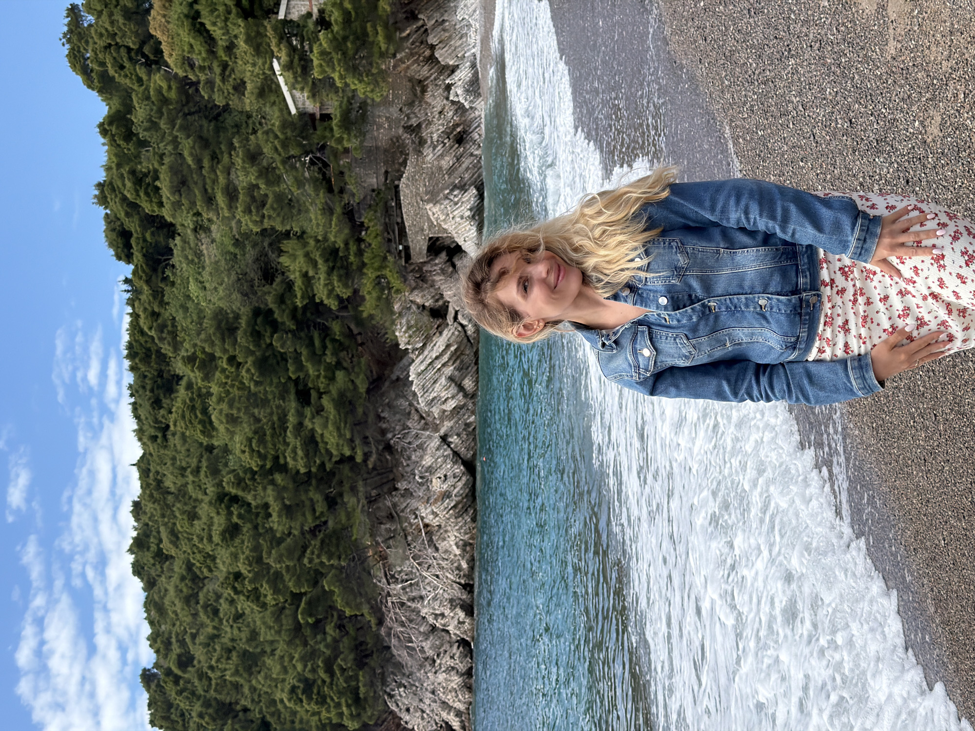 A woman with blonde hair stands on a pebble beach in Petrovac centar, Montenegro. She wears a denim jacket over a white dress with a small red floral print. The beach is washed by clear turquoise water with white foamy waves. Behind her to the left, a steep rocky cliff rises, covered with dense green vegetation. The sky is blue with scattered white clouds.