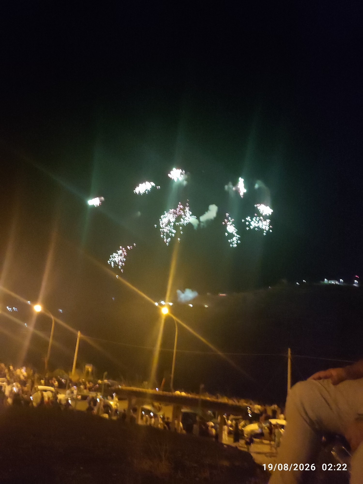 The image depicts a nighttime scene with fireworks exploding in the sky. A crowd of people is gathered below, observing the display. Streetlights illuminate the area, casting a warm glow. In the foreground, a person's legs and torso are visible, suggesting they are sitting or standing, facing away from the camera. The date and time stamp "19/08/2026 02:22" is overlaid on the lower right corner of the image. The overall atmosphere is festive, with the fireworks creating a dynamic and colorful spectacle against the dark sky.