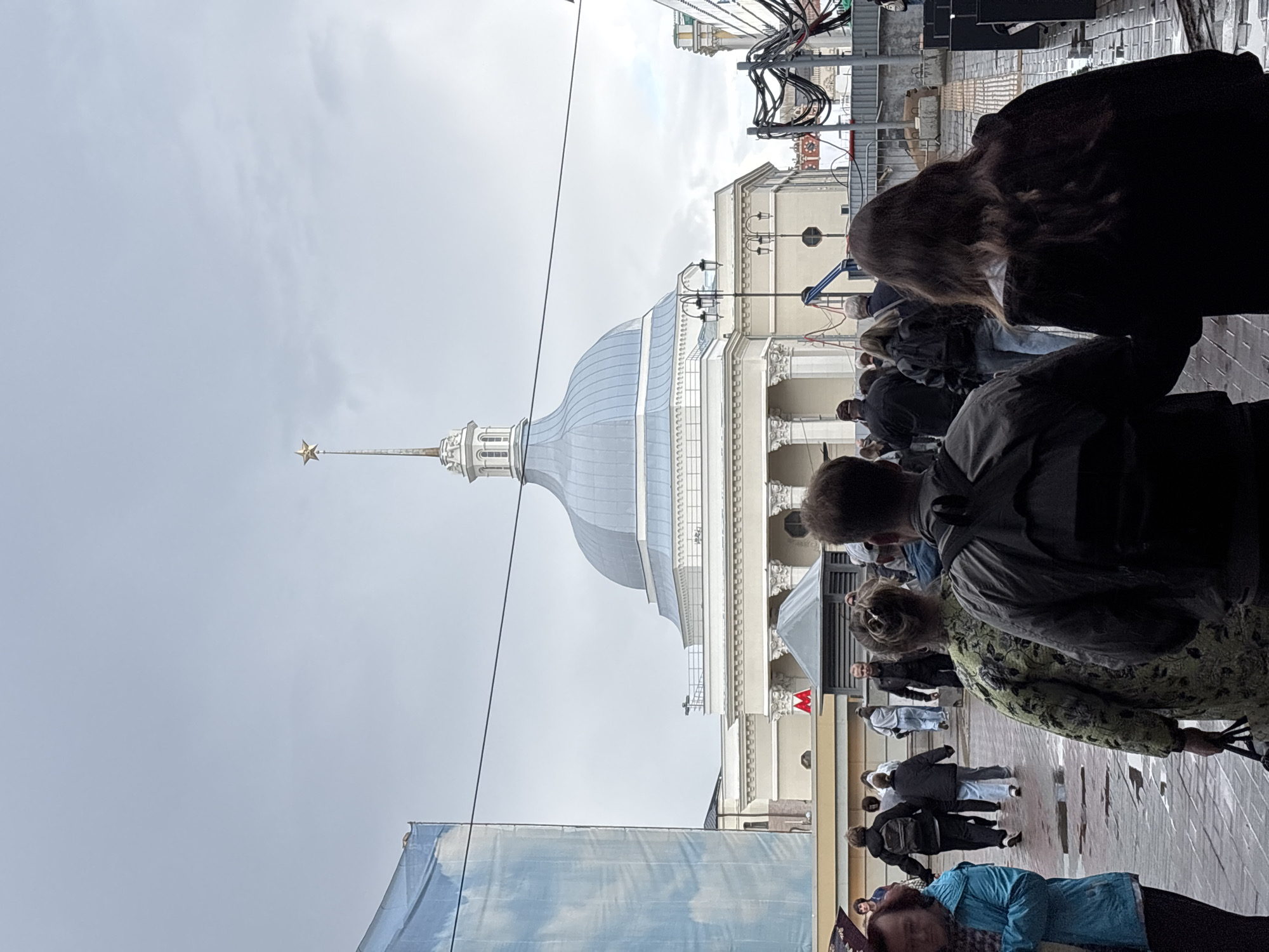 The image captures a street scene in Moscow, Russia, with a prominent building featuring a dome topped with a star, likely a significant architectural landmark. A crowd of people is gathered in front of the building, suggesting an event or a tourist attraction. The weather appears overcast, with a bright sky indicating it might be daytime, possibly late morning or afternoon. The ground is paved with cobblestones, and there's a sense of urban activity. Some individuals are facing the building, possibly observing or participating in something. The presence of what looks like a metro sign with a red "M" hints at the proximity of a subway station. The overall mood seems neutral, with people engaged in everyday activities or sightseeing. The image is oriented vertically, requiring a rotation to view it correctly.
