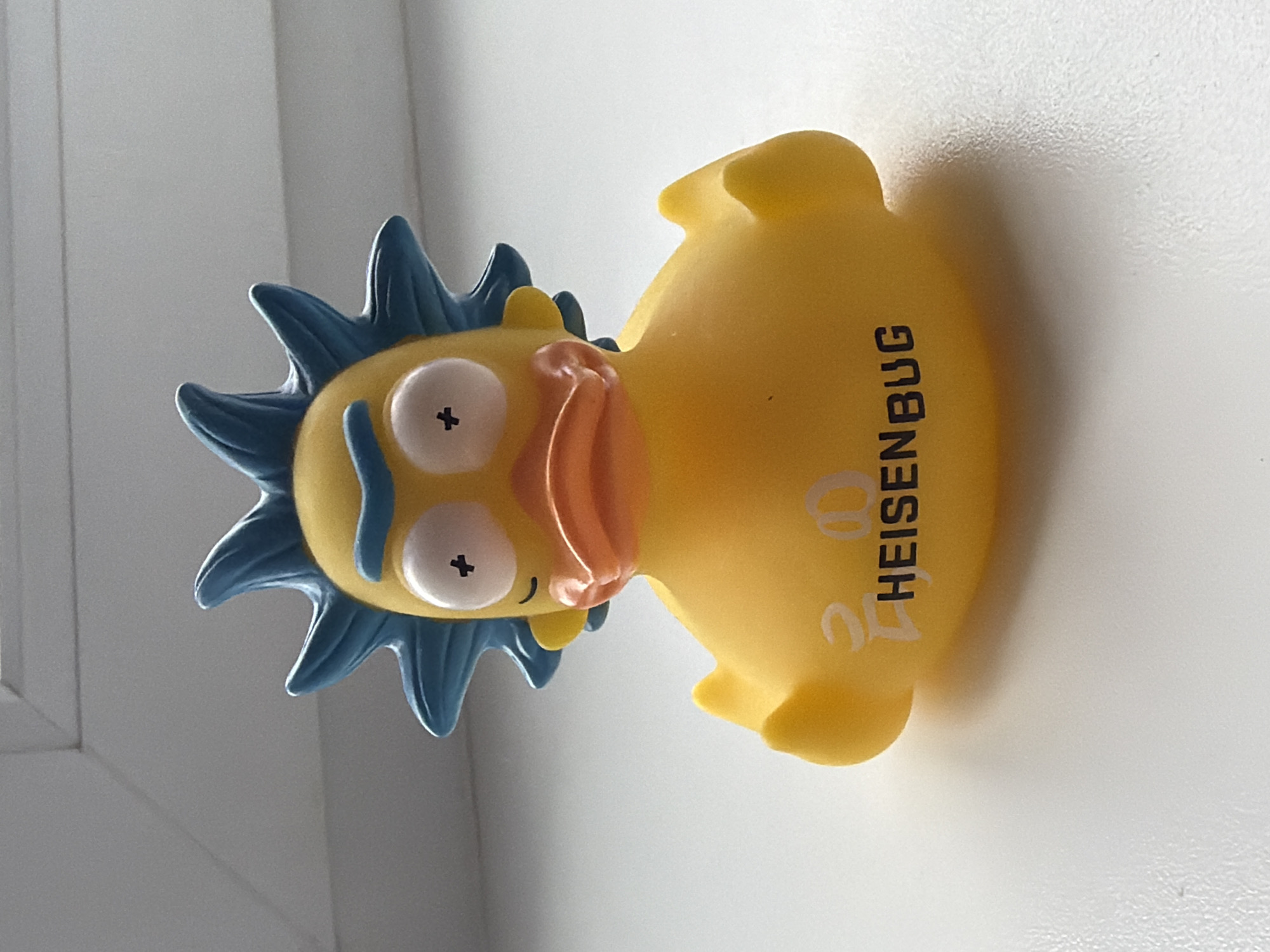 A yellow rubber duck toy, modeled after the character Rick Sanchez from the animated TV series "Rick and Morty," is lying on its side. The duck features Rick's signature blue spiky hair, bulging eyes with X marks, and a small orange beak. The word "HEISENBUG" is printed vertically on the duck's side in black ink, with two small, stacked circles above the word. The duck is positioned against a white background, suggesting it is indoors near a wall or window frame. No people or other prominent objects are visible in the scene. There is no discernible evidence to suggest the duck is in Chaikovsky, Russia.