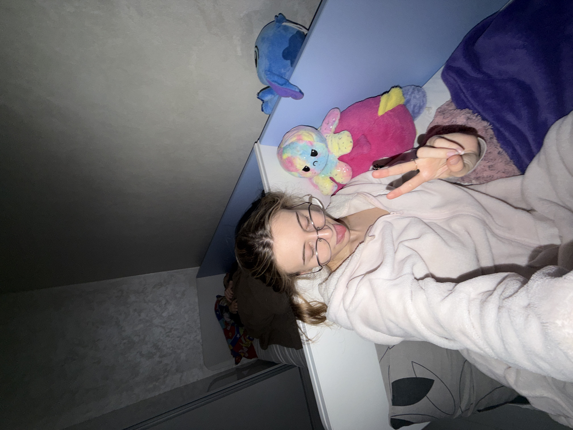 A young woman with glasses is lying in bed. She is wearing a light pink, fluffy robe and making a peace sign with her fingers. Beside her on the bed are two stuffed animals: a blue Stitch character and a pink octopus. Above her bed is a shelf with a blue finish. To the left, a wall is visible with a textured pattern. In the background, a glimpse of another bed with colorful pillows is visible. There is no evidence suggesting this image was taken in the city of Plovdiv, Bulgaria.