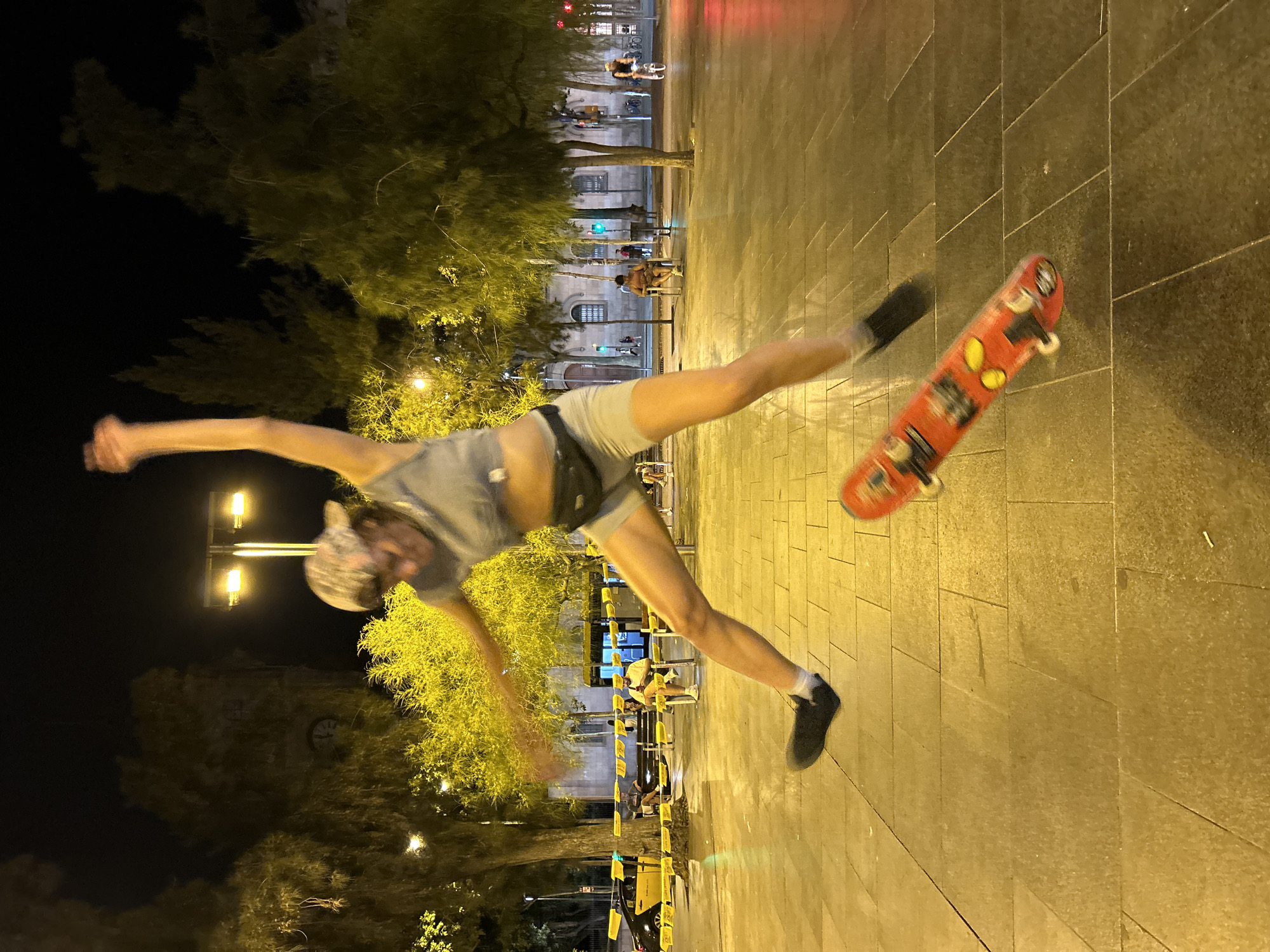 A person is captured mid-air, jumping over a skateboard in a public square at night. The skateboard, adorned with colorful stickers, is positioned beneath the person's airborne body. The individual, wearing a grey crop top, grey shorts, a black fanny pack, and black shoes with white socks, is in the process of performing a trick. The background reveals a nighttime cityscape, likely in Barcelona, Spain, featuring trees illuminated by yellow streetlights, buildings with windows, and street lamps. Other people are visible in the distance, some seated on benches and others walking or cycling, suggesting a lively urban environment. The ground is paved with large, square tiles, reflecting the ambient light.