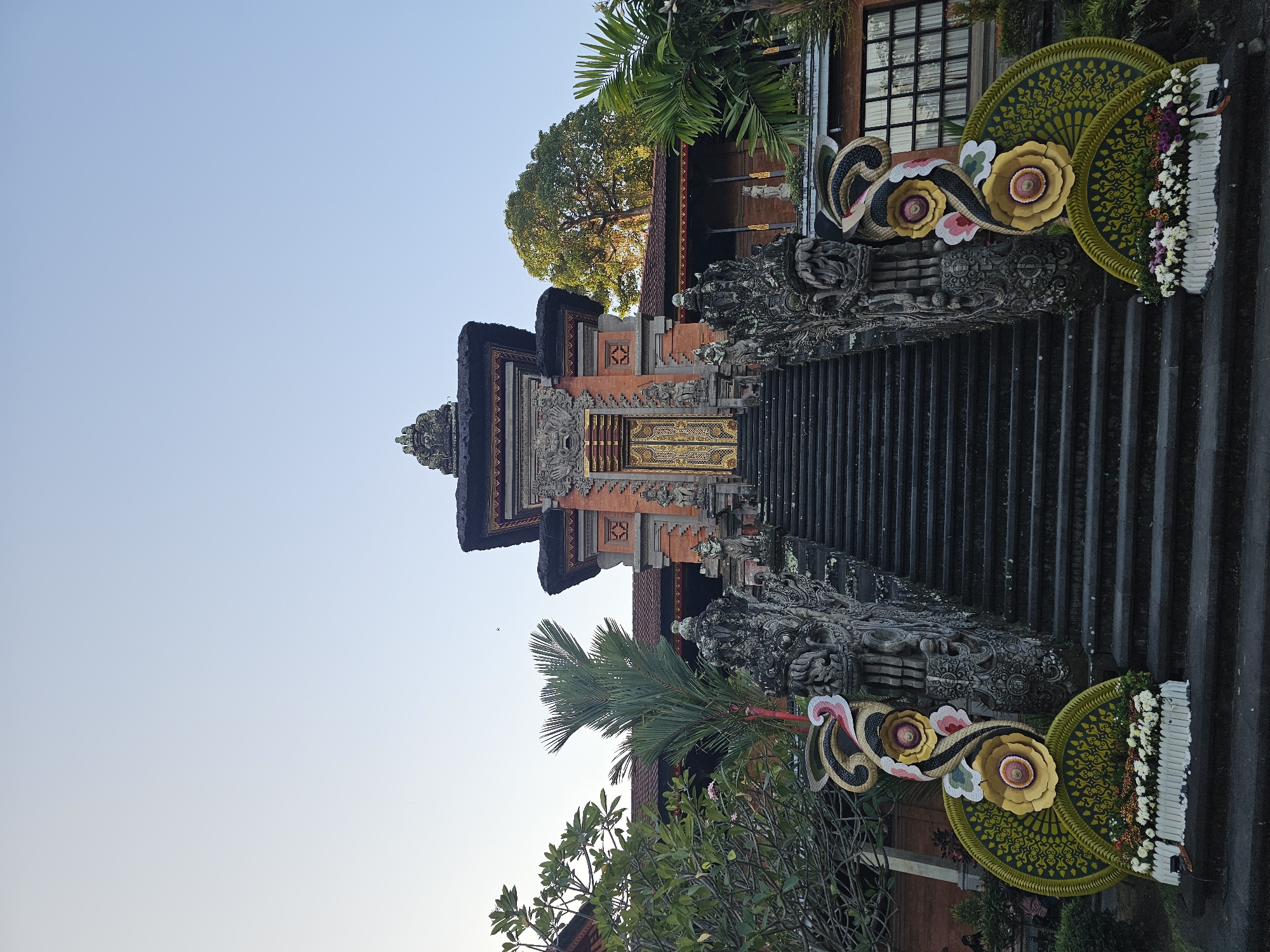 The image shows the ornate entrance to a temple, likely in Denpasar, Indonesia, given the architectural style. A wide, dark stone staircase ascends towards a intricately carved stone gateway adorned with golden doors. The gateway is topped with a multi-tiered roof structure with dark thatch and decorative stonework. Flanking the staircase are elaborately carved stone balustrades, featuring mythical creatures and intricate patterns. On either side of the base of the stairs, large, colorful floral arrangements are displayed, one with large yellow and pink flowers set against a green patterned backdrop, and the other with a white border filled with white and purple flowers. Lush green foliage, including palm trees and leafy bushes, surrounds the temple structure, adding to the tropical ambiance. The sky above is clear and pale blue. No people are clearly visible in the image. The scene suggests a place of cultural or religious significance, possibly decorated for a festival or ceremony.