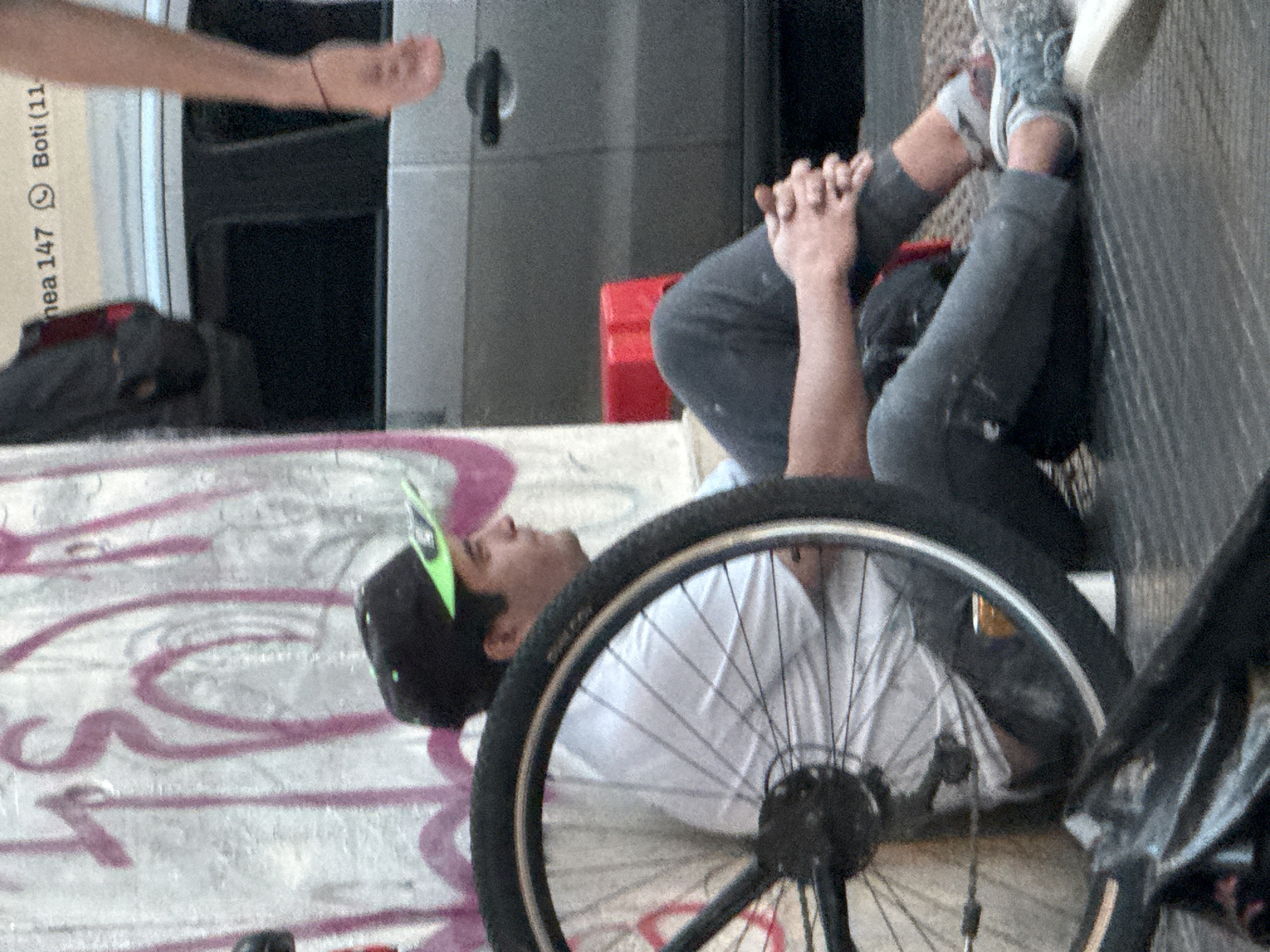 The image depicts an outdoor urban scene, likely in Buenos Aires, Argentina. A person, appearing to be a young man, is lying on their back on a concrete surface heavily marked with purple and pink graffiti. He is wearing a white t-shirt, dark pants, and sneakers, with a black cap featuring green accents. His knees are bent, and his hands are clasped over them, suggesting a moment of rest or relaxation. His face is visible, looking upwards.
In the foreground, a prominent black bicycle wheel with silver spokes and part of its frame are visible, positioned close to the resting individual. In the upper left background, another person's arm and hand extend into the frame. Further in the background, there is a plain gray surface, possibly a door or part of a building, and a red object that resembles a crate or box.
Visible text on a light-colored vertical surface in the upper left corner includes "enea 147" and "Boti (11", accompanied by a WhatsApp logo. The scene conveys a candid moment of leisure or contemplation in a bustling urban environment, characterized by street art and everyday elements.
