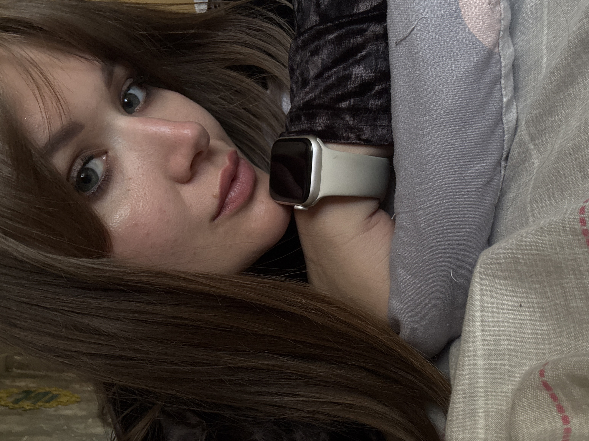 A young woman with light eyes and brown hair is lying down, with a smartwatch on her wrist. Her head is tilted towards the camera. She is partially covered by bed linens and a textured blanket or garment. The exact location in the city of Chaikovsky, Russia is not discernible from the image.
