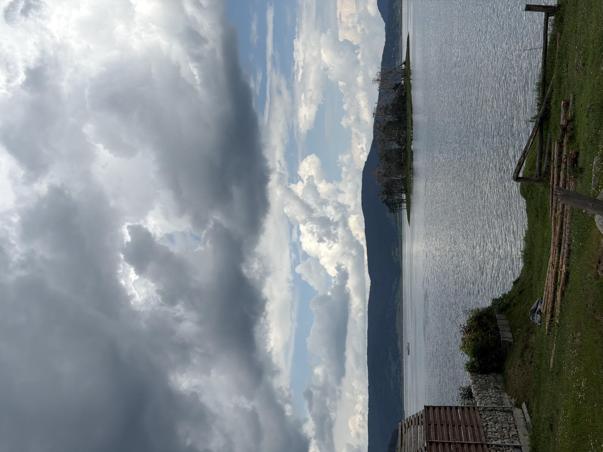This wide-angle landscape photograph depicts a scene at the Batak Reservoir, located near the town of Цигов чарк, Bulgaria, under a dynamic sky. The upper two-thirds of the image are dominated by a heavy, turbulent cloud cover, transitioning from dark grey, dense cumulus clouds in the upper left to brighter white clouds and intermittent patches of blue sky towards the upper right and central horizon.
The mid-ground features a large body of water, its surface exhibiting numerous small ripples, reflecting the subdued light from the sky. Approximately one-third across from the left edge and halfway down from the top, a small, light-colored boat carrying three to four indistinct human figures navigates the water. Towards the right side of the water body, in the mid-ground, a small, irregularly shaped island is present, densely covered with dark, deciduous trees, some appearing sparse or bare-branched. Beyond the water, a continuous line of dark, forested hills or mountains extends across the horizon, forming the distant background.
In the immediate foreground on the left, a partial view of a building is visible. It features a facade constructed with horizontal wooden slats above a stone-stacked wall base. Adjacent to the building, a small area of green grass slopes gently towards the water. On this grassy incline, a pile of approximately ten cut logs of varying lengths is laid out horizontally. To the right of the logs, a rustic wooden fence, appearing partially broken or unfinished, extends towards the water's edge. A dark, possibly bundled object, resembling a tarp or folded fabric, lies on the grass near the logs. Green bushes and other foliage are present between the building and the water, partially obscuring the immediate shoreline. No clear human activity or interaction is depicted beyond the distant boat.