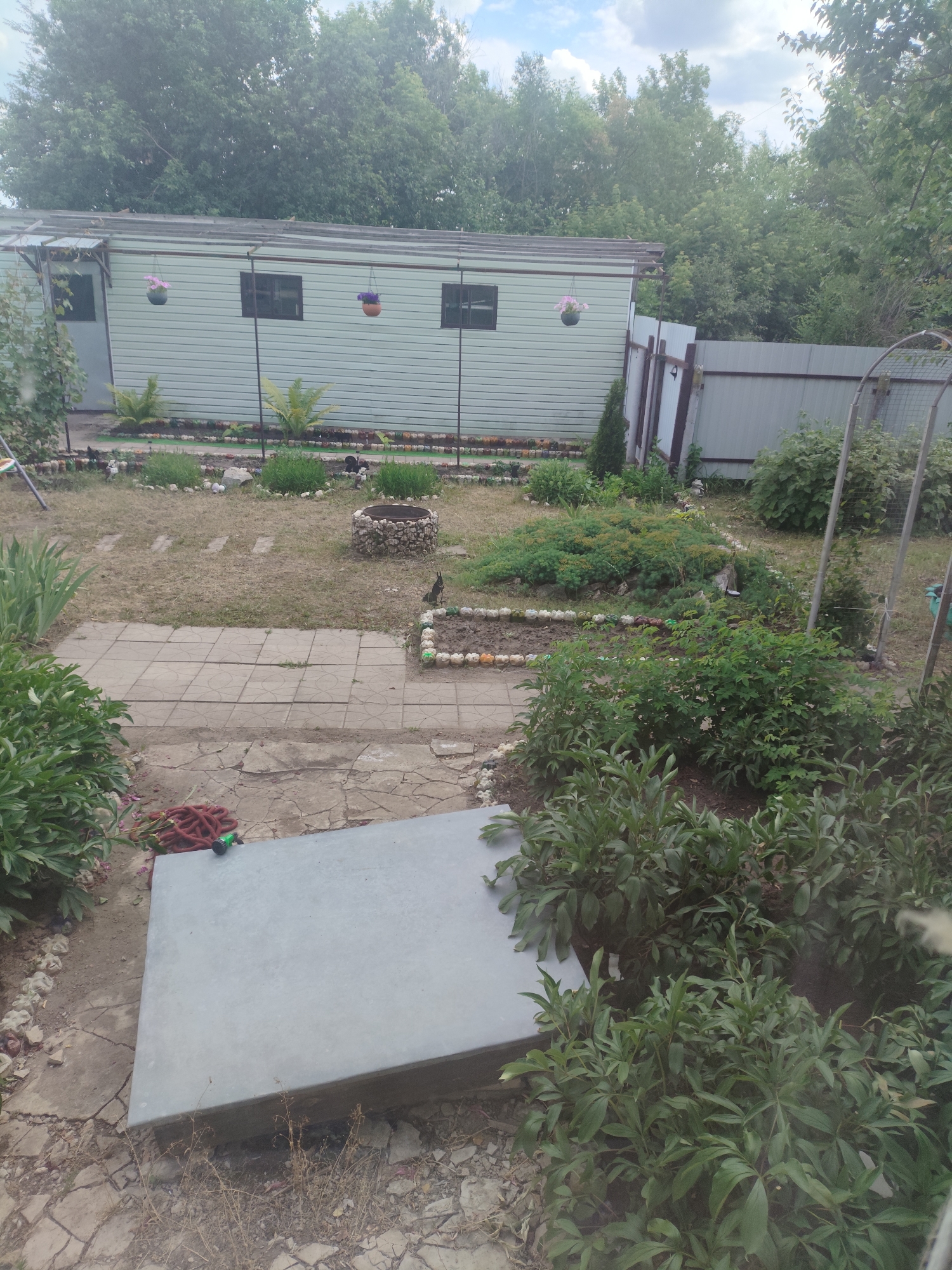 This is an outdoor scene of a backyard in Aleksikovskii, Russia. The image captures a slightly overcast day, suggested by the diffused light and cloudy sky. A light green, rectangular building, possibly a shed or workshop, stands prominently in the background, with two windows visible. In front of the building, several potted plants are hung, adding a touch of color to the scene.
The yard itself is a mix of dry grass, paved pathways made of square tiles, and various garden beds. Some beds are outlined with small stones and contain low-lying green plants. A circular stone structure, possibly a small fire pit or decorative element, is visible in the mid-ground. Further into the yard, a metal fence marks the property line, with dense trees and foliage beyond.
In the foreground, a large, flat, grey concrete slab rests on the ground, partially covered by some dry vegetation and a red garden hose with a green nozzle. Lush green bushes and plants are positioned to the left and right of this slab, indicating a well-maintained garden area. The overall impression is that of a functional, lived-in backyard, with elements of both practical use and decorative gardening. There is no visible text in the image.