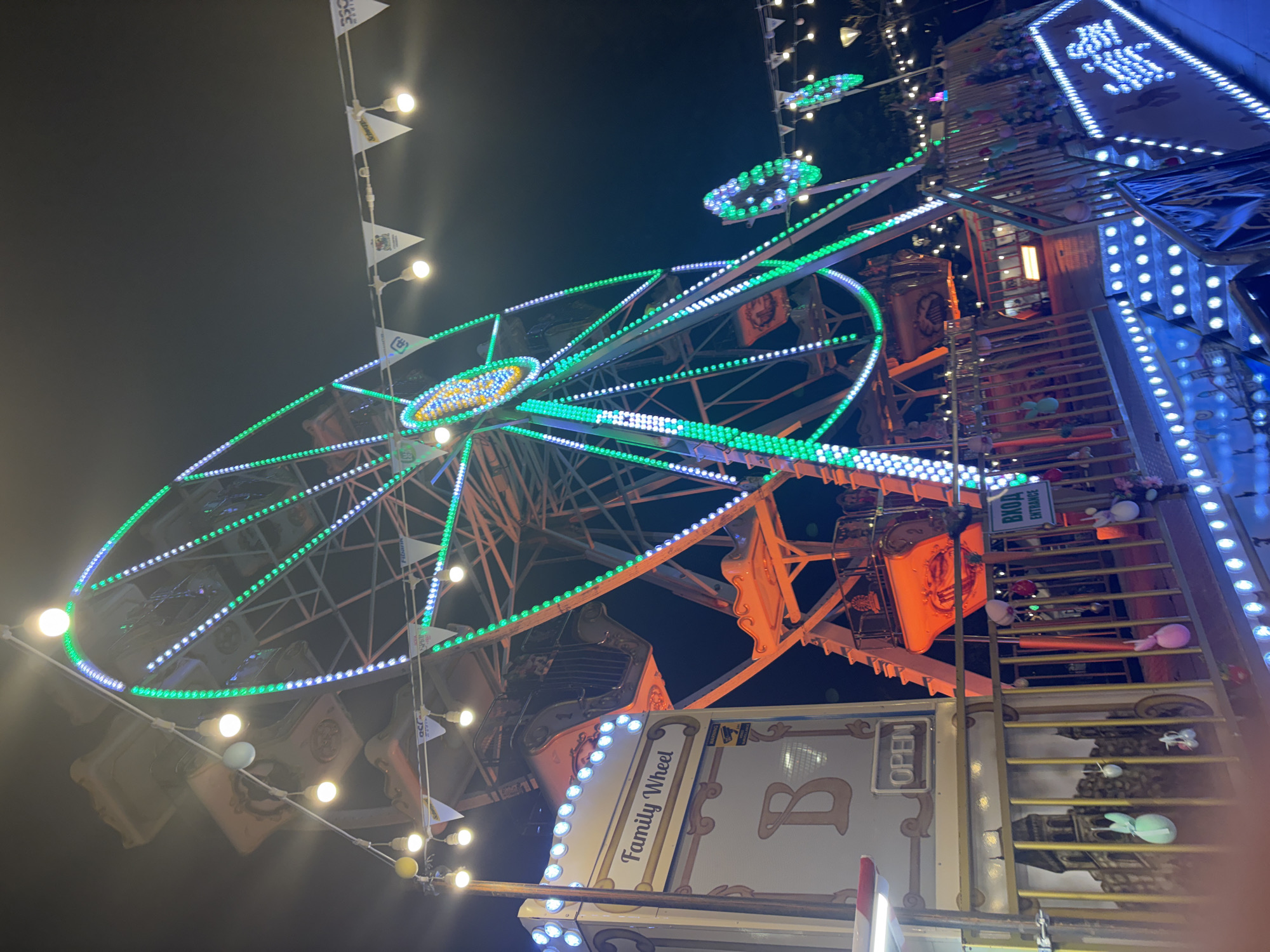 This media file captures a vibrant night scene at what appears to be a fairground or carnival in Sofia, Bulgaria. The central subject is a large Ferris wheel, brilliantly illuminated with rows of green, white, and blue LED lights that trace its circular structure and spokes. Its gondolas are visible in an orange-brown hue.
To the right of the Ferris wheel, a tall, multi-level structure extends upwards, also adorned with numerous white and blue lights, resembling a show booth or an intricate entrance to an attraction. A staircase or ramp leads up this structure, and various plush toys, including bunnies, are arranged on its steps and railings, suggesting they might be prizes or decorations.
The scene is captured at night, with the dark sky serving as a backdrop to the dazzling artificial lights. White spherical lights are strung on bunting across the top of the image, contributing to the festive ambiance. No people are clearly visible on the Ferris wheel or in the immediate foreground, allowing the focus to remain on the illuminated rides.
Several pieces of text are visible, providing specific details. A sign near the bottom right clearly reads "Family Wheel," with a large, stylized letter "B" next to it. Another sign, positioned vertically above this, displays "OPEN." Further up, on a green sign near the entrance to the structure, both Cyrillic and English text is visible: "ВХОД" and "ENTRANCE," respectively. Logos that appear to be "Fitcan" or similar are faintly discernible on some of the small flags decorating the scene. The abundance of colorful lights and the presence of amusement rides create a lively and entertaining atmosphere characteristic of a nighttime carnival event.