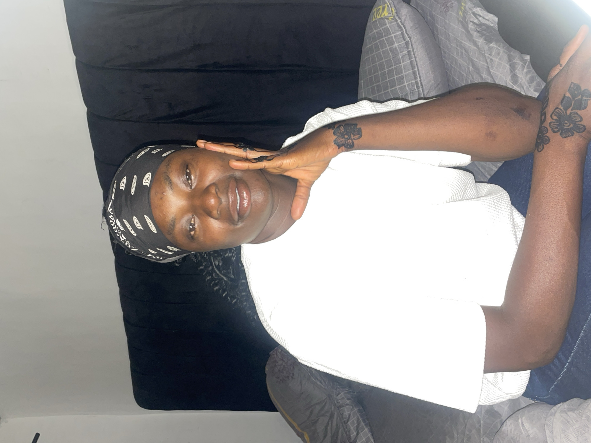 A young person with dark skin is captured in a portrait, appearing to be lying down or reclining indoors, with the image rotated 90 degrees counter-clockwise. They gaze directly at the camera with a gentle smile or smirk. The individual wears a black head covering with a white pattern, a plain white long-sleeved top, and blue jeans. Both hands are prominently adorned with intricate black floral henna or temporary tattoo designs; the left hand is raised towards the face, showcasing detailed patterns on the fingers and palm, while similar designs are visible on the back of the right hand.
The background consists of a dark, possibly black, upholstered headboard or backrest behind the person. To the right, a gray and white patterned pillow is visible, featuring some indistinct yellow text that is illegible. A plain light-colored wall forms part of the setting. The scene conveys a relaxed and calm atmosphere, likely an indoor environment such as a bedroom or living room in Jos, Nigeria. The lighting suggests an indoor setting.