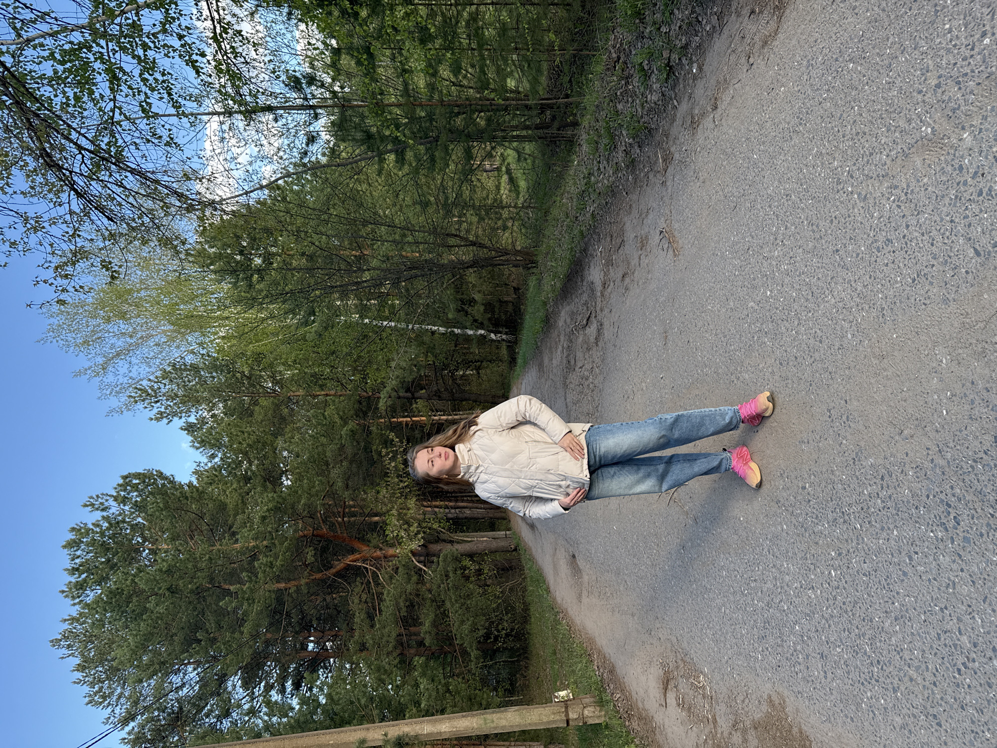A full-length shot shows a young woman standing on an asphalt road in a wooded area. She is wearing light blue jeans, a beige jacket, and pink sneakers. The road stretches away from the viewer. Trees line one side of the road. The sky is visible through the trees.