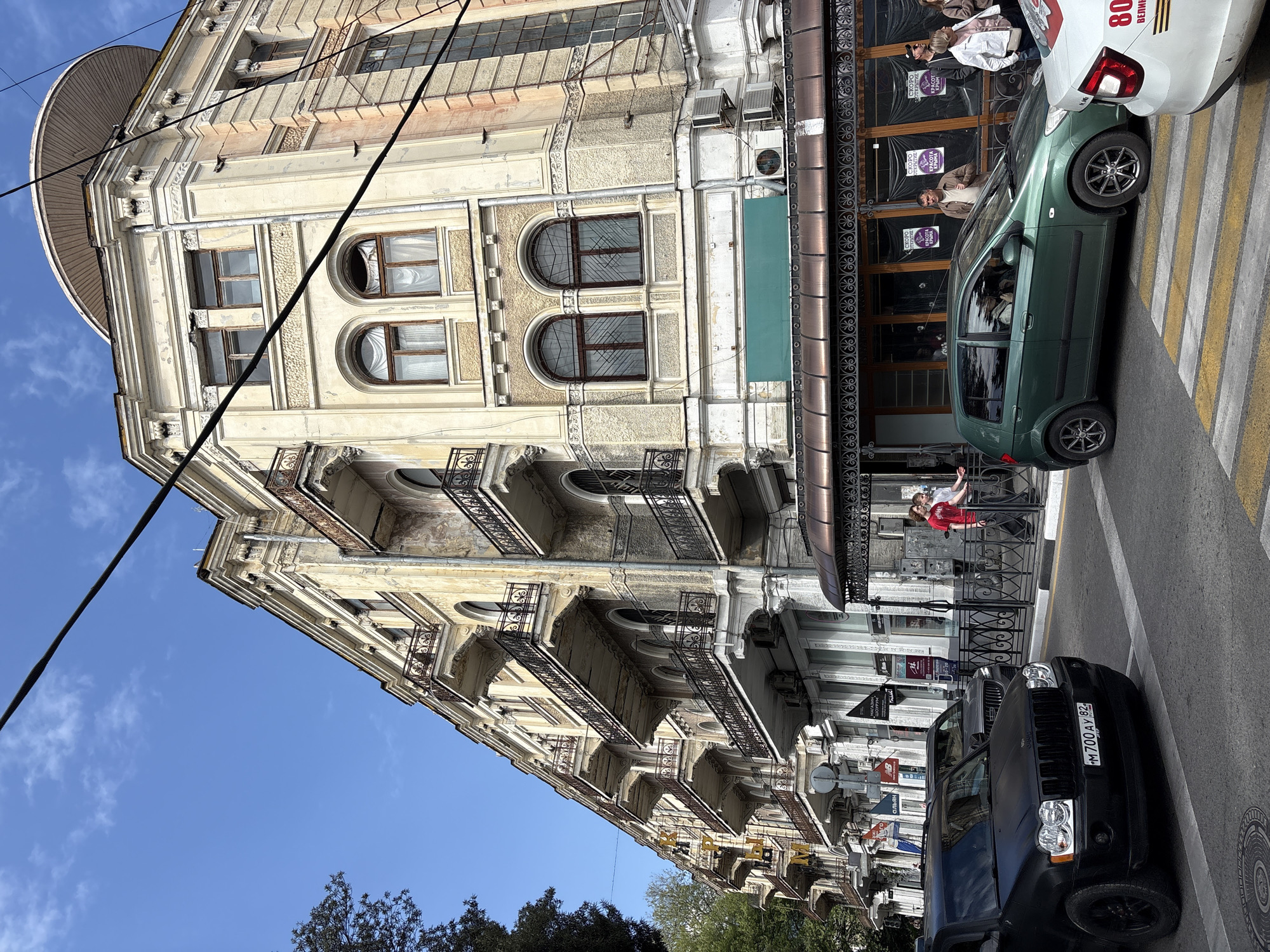 The image shows a street scene in город Ялта, Ukraine, featuring a tall, ornate building with multiple floors and balconies. The building has arched windows and decorative stonework. Power lines run across the upper left of the frame, contrasting with the architecture.
On the street level, there is a storefront with a dark awning and patterned glass doors. A green compact car is parked on the crosswalk. To the right is a white vehicle. A black SUV is parked in front of the building with a license plate visible.
Two people, one wearing a red shirt, stand near the storefront, possibly interacting with the storefront or each other. The environment is sunny, with blue sky visible above the building and in the gaps between the trees below. Other parked cars and street signs are visible further down the street.