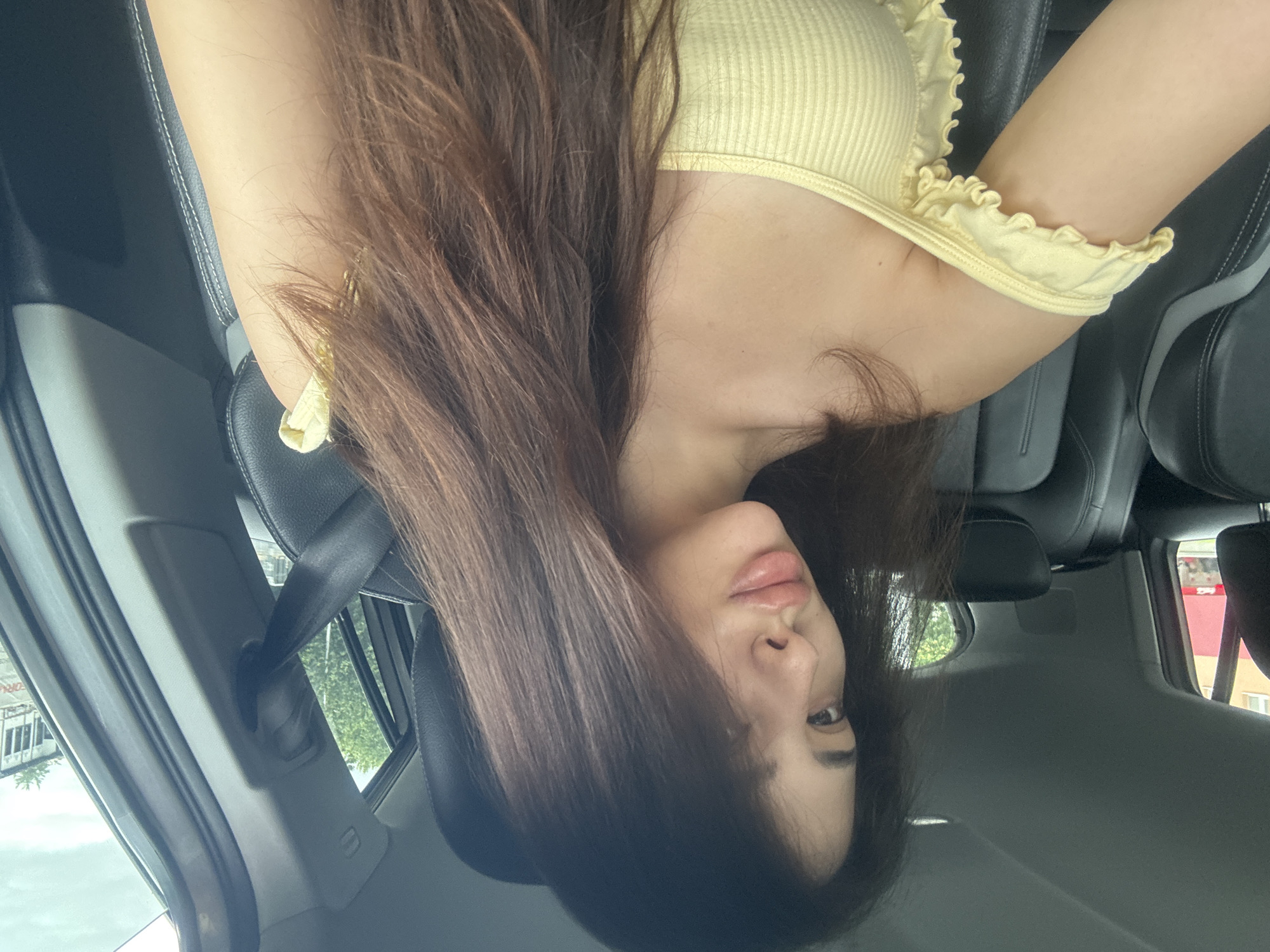 A young woman is captured in a close-up, rotated image, seated in the passenger seat of a car. She has long, dark brown hair that falls over her shoulders and chest, and fair skin with full lips. Her head is tilted, and she looks towards the camera with a neutral, relaxed expression. She is wearing a light yellow, ribbed crop top featuring ruffled detailing along the neckline and straps.
The car's interior is visible, showing black leather seats with white stitching and a black seatbelt across her chest. Through the car window, bright daylight illuminates an outdoor scene. Green foliage from trees and several buildings can be observed, indicating an urban or semi-urban setting. On one building in the background, faint, red, and largely indistinct lettering, possibly "PR...ICE," is barely visible. The lighting suggests the photo was taken during the daytime. The context provided indicates the location is Plovdiv, Bulgaria.