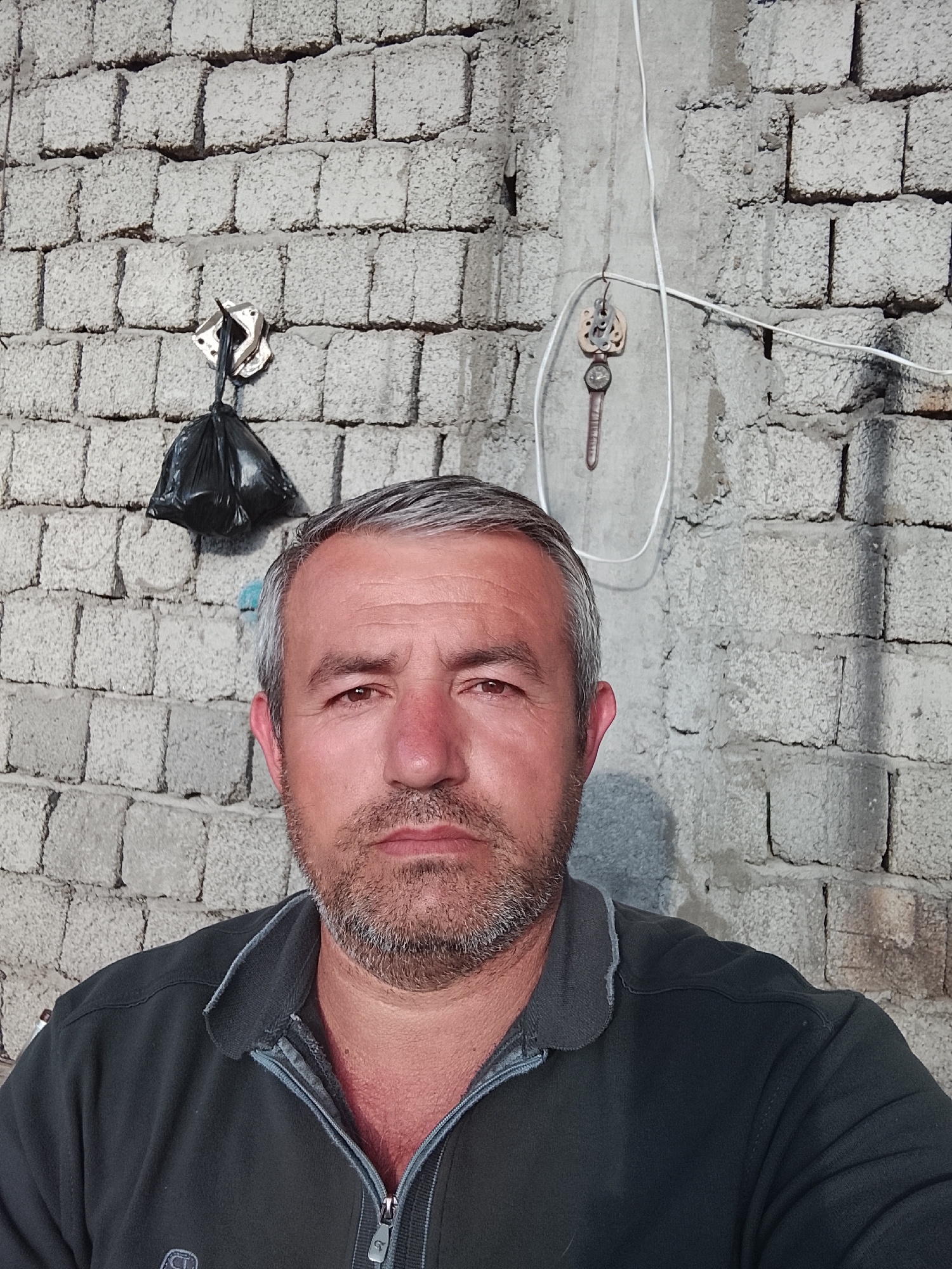 A middle-aged man with graying hair and a short beard is standing in front of a brick wall. A black plastic bag hangs on a metal hook to the left of his head. A decorative item resembling a watch and gears is hanging on the wall above his right shoulder. A white electrical wire is also visible on the wall. The man is wearing a dark-colored zip-up shirt. The location is potentially in Sabir Rakhimov, Uzbekistan, given the request for the location. The background appears to be outdoors.