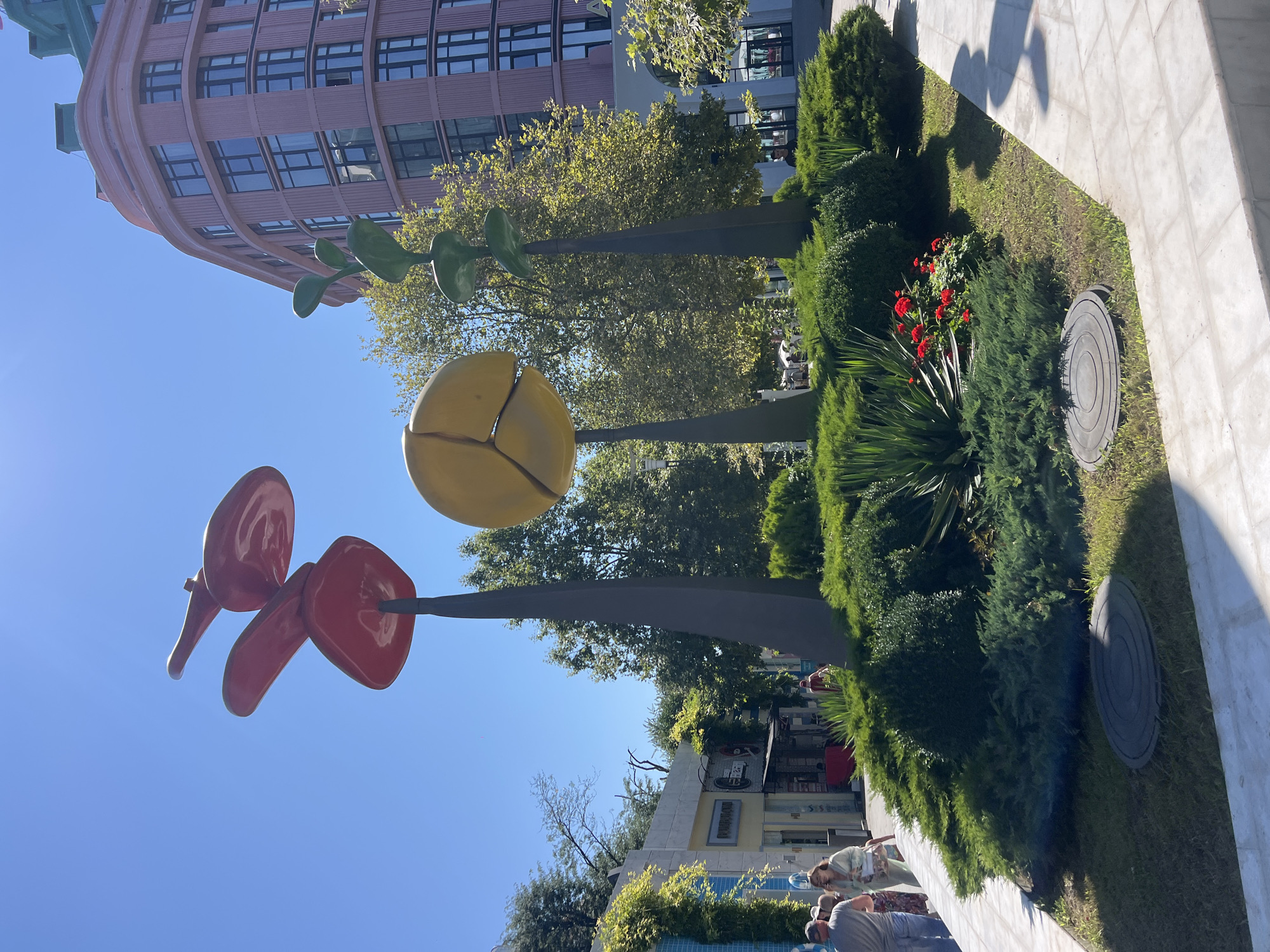 In Odesa, Ukraine, a sunny outdoor scene features large, colorful abstract sculptures resembling oversized plants or flowers. The sculptures are situated in a landscaped area with lush green bushes and some red flowers. Behind the sculptures, a modern pink building with multiple windows curves around a corner. To the left of the sculptures, there are storefronts with awnings and people walking by on a sidewalk. The sky is clear and blue.