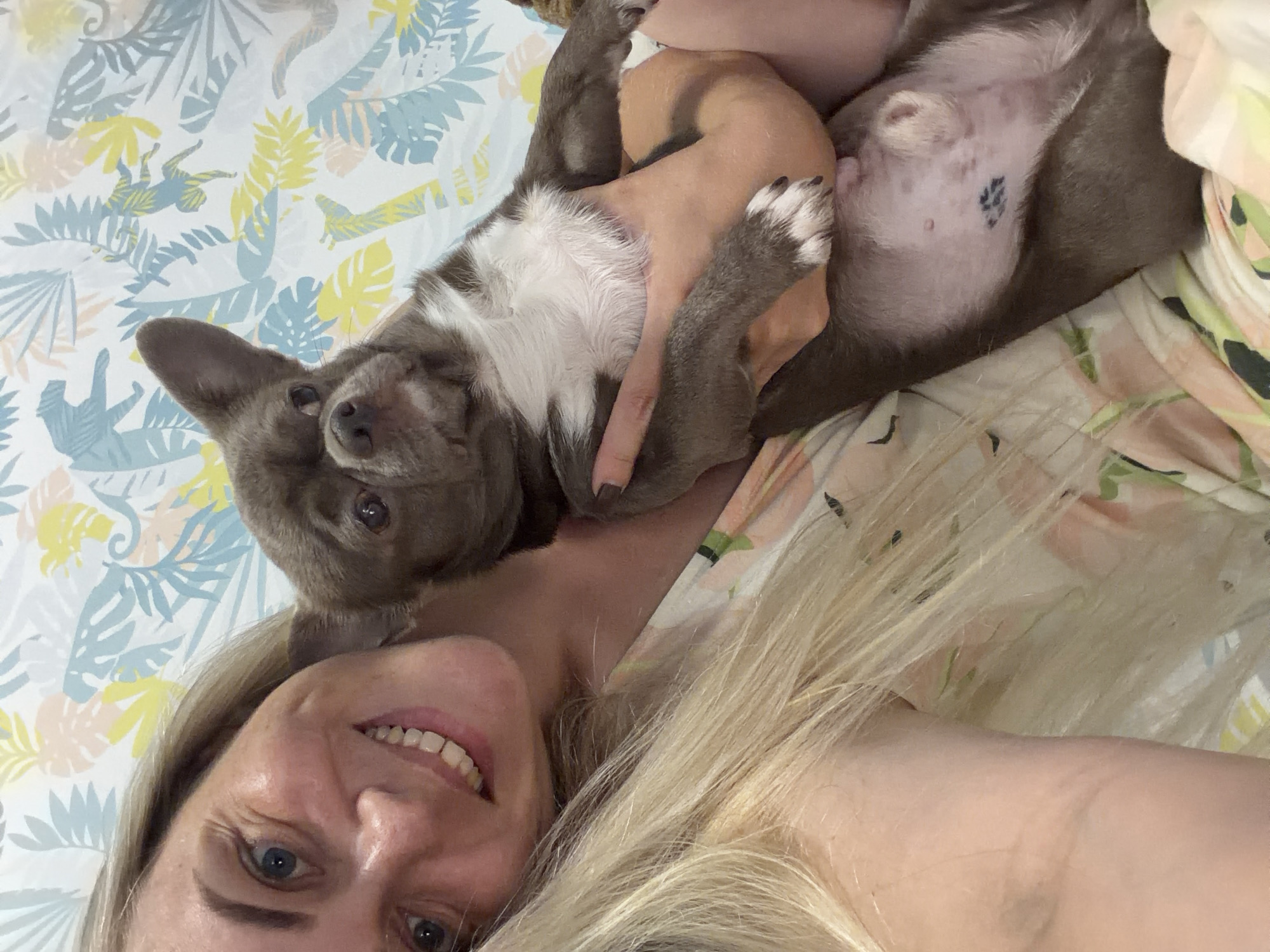 The image shows a selfie taken from a high-angle perspective. The main subjects are a woman and a young dog, likely a puppy, lying together on a bed. The woman has blonde hair and is smiling, looking towards the camera. Her face is in the foreground and is slightly blurred. The puppy is held in her arms, with its belly exposed. It appears to be a grey and white breed with dark eyes. A small, dark paw print tattoo is visible on the puppy's belly. The background features a colorful tropical-themed fabric, possibly a bedsheet or duvet cover, with patterns of leaves and stylized figures in shades of blue, yellow, and pink. The lighting suggests daytime. The overall mood is intimate and affectionate, capturing a moment of bonding between the woman and her pet. The location context of Votkinsk, Russia, is provided but not visually evident in the image. There is no visible text in the image.
