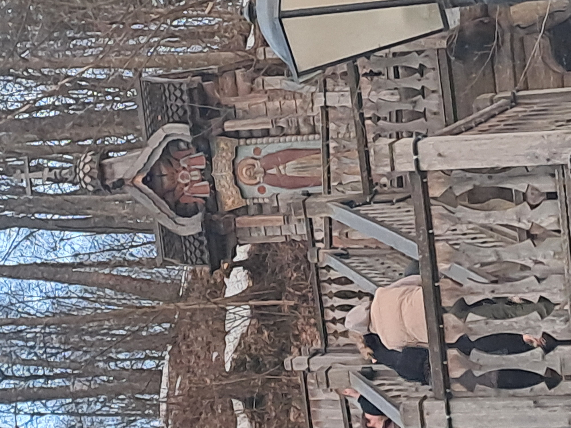 A tilted medium shot shows a wooden structure next to a leafless tree in Vzglyadnevo, Russia. The structure features intricate carvings and a painting of a figure with a halo, possibly a religious icon. A partial view of a person in dark clothing and a light-colored top is visible on the structure. A lamppost sits atop the structure. The environment is mostly natural with bare trees and a hint of sky in the background.