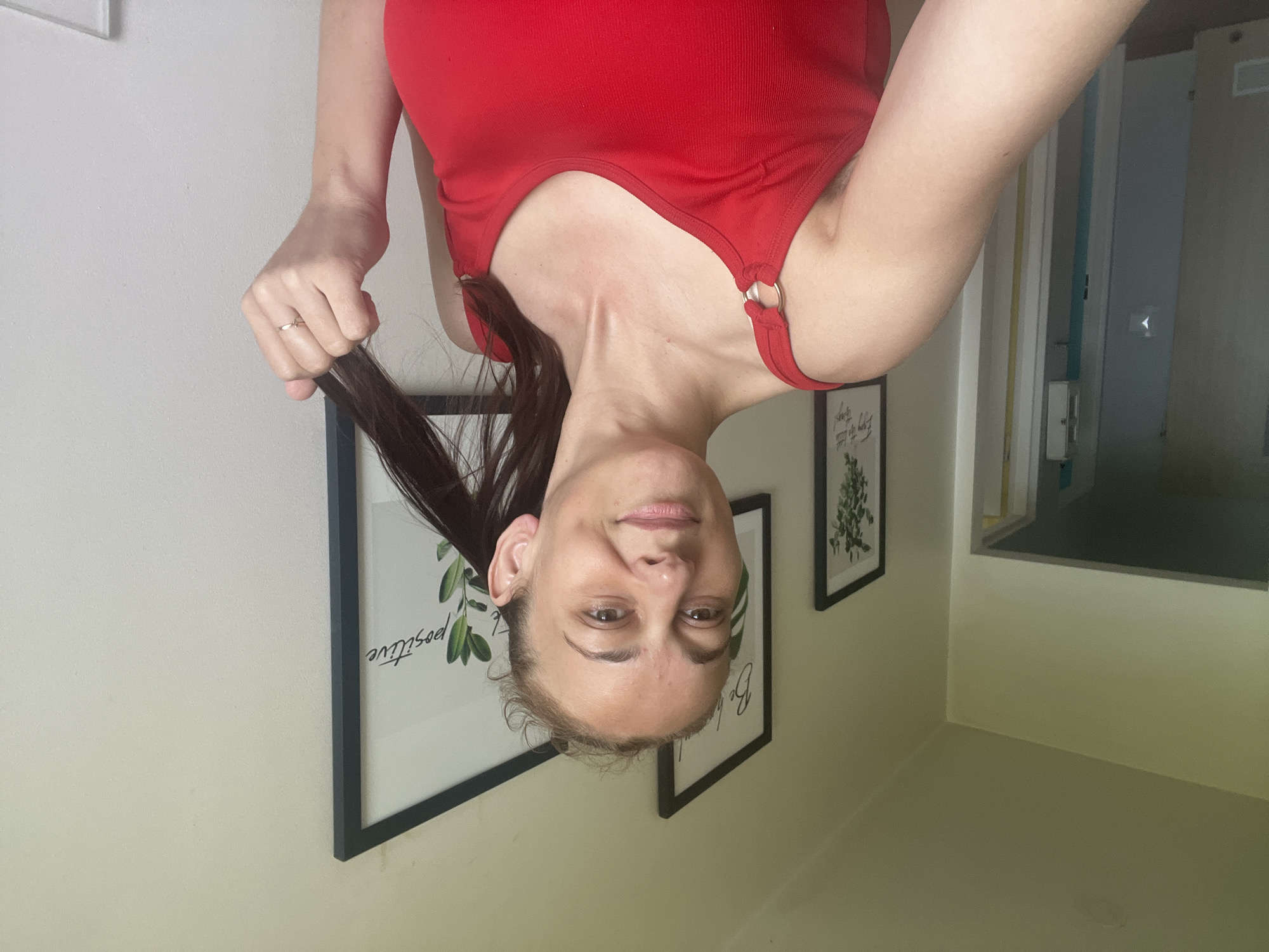 A woman with light skin and brown hair is in a room, holding a lock of her hair with her right hand. She is wearing a red tank top. In the background, there are two framed pictures on the wall and a doorway. The lighting suggests it is daytime.