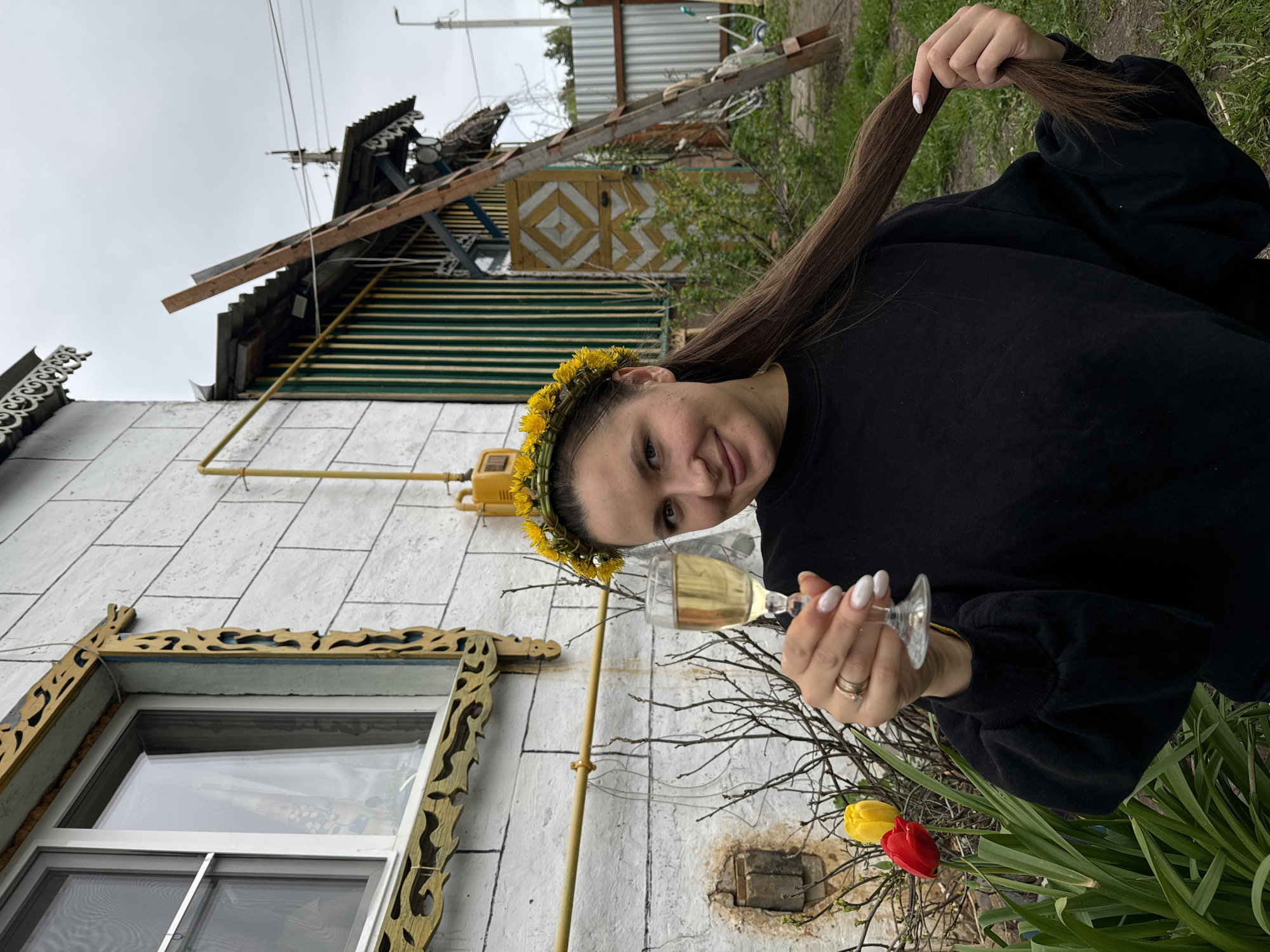 A young woman wearing a dandelion crown and holding a glass of sparkling wine or champagne poses outdoors in Красный Маяк, Russia. She wears a black sweatshirt and holds a long strand of brown hair in her right hand. She smiles at the camera, showing a ring on her left ring finger. A building with white siding and yellow trim is behind her, including a window with ornate wooden detailing. Yellow gas pipes run along the side of the building. Tulips, one red and one yellow, are at the bottom right of the frame. The environment suggests a rural setting with overcast skies.