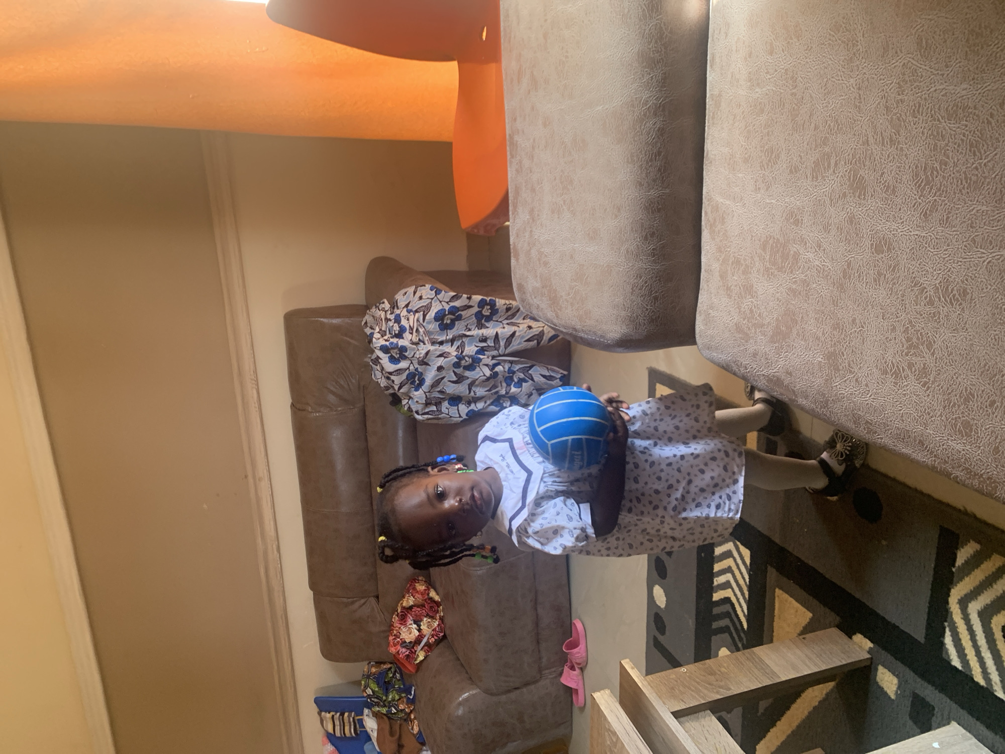 In Gura Top, Nigeria, a young Black girl stands in a living room holding a blue ball. She wears a white dress with a subtle pattern and dark sandals. The room contains a brown leather couch with patterned fabric draped over it, a wooden table, and a patterned rug on the floor. An orange chair and a partially visible light-colored upholstered seat are also present. The lighting suggests it is daytime, with natural light coming from the right side of the frame, illuminating the scene.