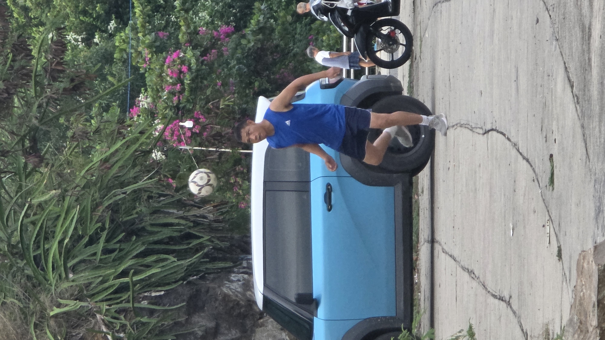 The image depicts an outdoor scene, likely in Bac Nha Trang Ward, Vietnam, captured during daylight with soft, diffused lighting.
In the central foreground, a young man is engaged in a dynamic activity. He wears a blue tank top with a white Adidas logo, dark shorts, white socks, and light-colored sneakers. He is positioned next to a light blue and white SUV, with his right leg raised as if kicking or balancing a white and black soccer ball. The ball is suspended in the air to his left, amidst a dense cluster of long-leafed green plants that occupy the left side of the frame.
The SUV is parked on a cracked concrete or asphalt surface, which forms a road or pathway extending across the middle and right side of the image. Further back on this path, two other individuals are visible; one stands near a black motorcycle, and another person in a white t-shirt and shorts. The background is filled with lush greenery, including tall trees and bushes adorned with bright pink flowering bougainvillea. A white light fixture, attached to a thin blue wire, is nestled among these pink blooms. The overall impression is one of casual outdoor activity within a verdant, semi-urban environment.