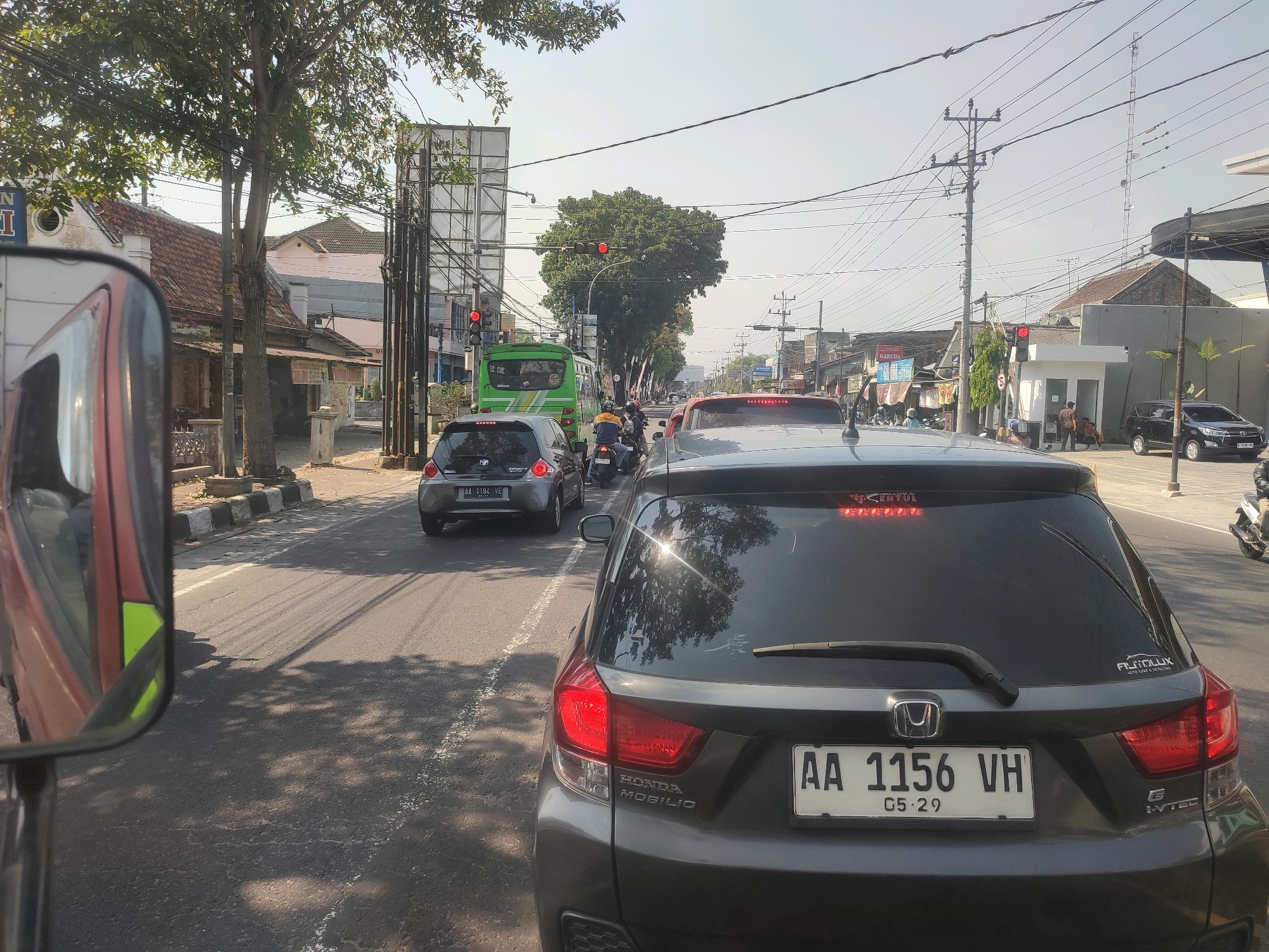 The image depicts a street scene in Magelang, Indonesia, during daylight. The perspective is from within a vehicle, looking forward and slightly to the right. Traffic is present, with several cars and motorcycles on the road. In the foreground, the rear of a dark-colored Honda Mobilio is visible, its license plate reading "AA 1156 VH" with an expiration date of "05.29". A green bus is also in traffic ahead of the Mobilio, along with other vehicles and motorcycles. The roadside features buildings, trees, utility poles with numerous wires, and traffic lights. One traffic light is red, indicating traffic is stopped. The environment appears to be a moderately busy urban or suburban area.