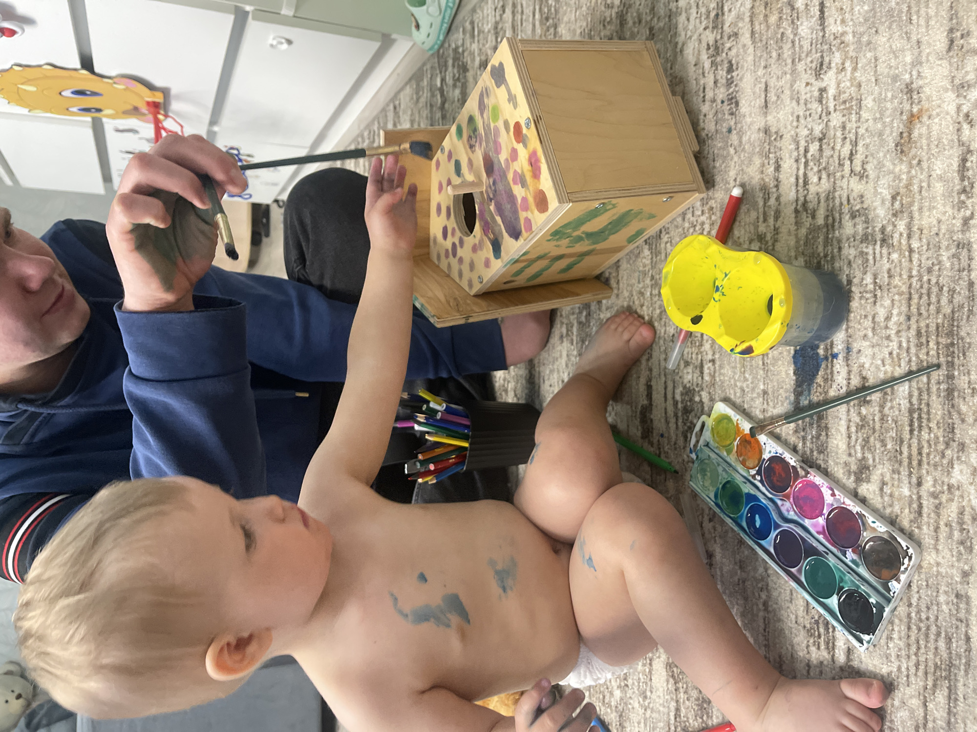 A person and a toddler are sitting on a carpeted floor next to a wooden birdhouse. The person, wearing a blue hoodie, holds a paintbrush in their right hand, which is covered in blue paint, and appears to be painting the birdhouse. The toddler, wearing a diaper and bare-chested, has blue paint smudged on their torso and limbs and is holding something small in their right hand. The birdhouse is partially painted with colorful spots and shapes. A yellow cup with two sections, a container of colored pencils, a palette of watercolors, and several brushes are scattered on the carpet around them. There is furniture visible in the background, including a white cabinet. The location is not discernible, and there is no information to indicate this is Izhevsk, Russia.