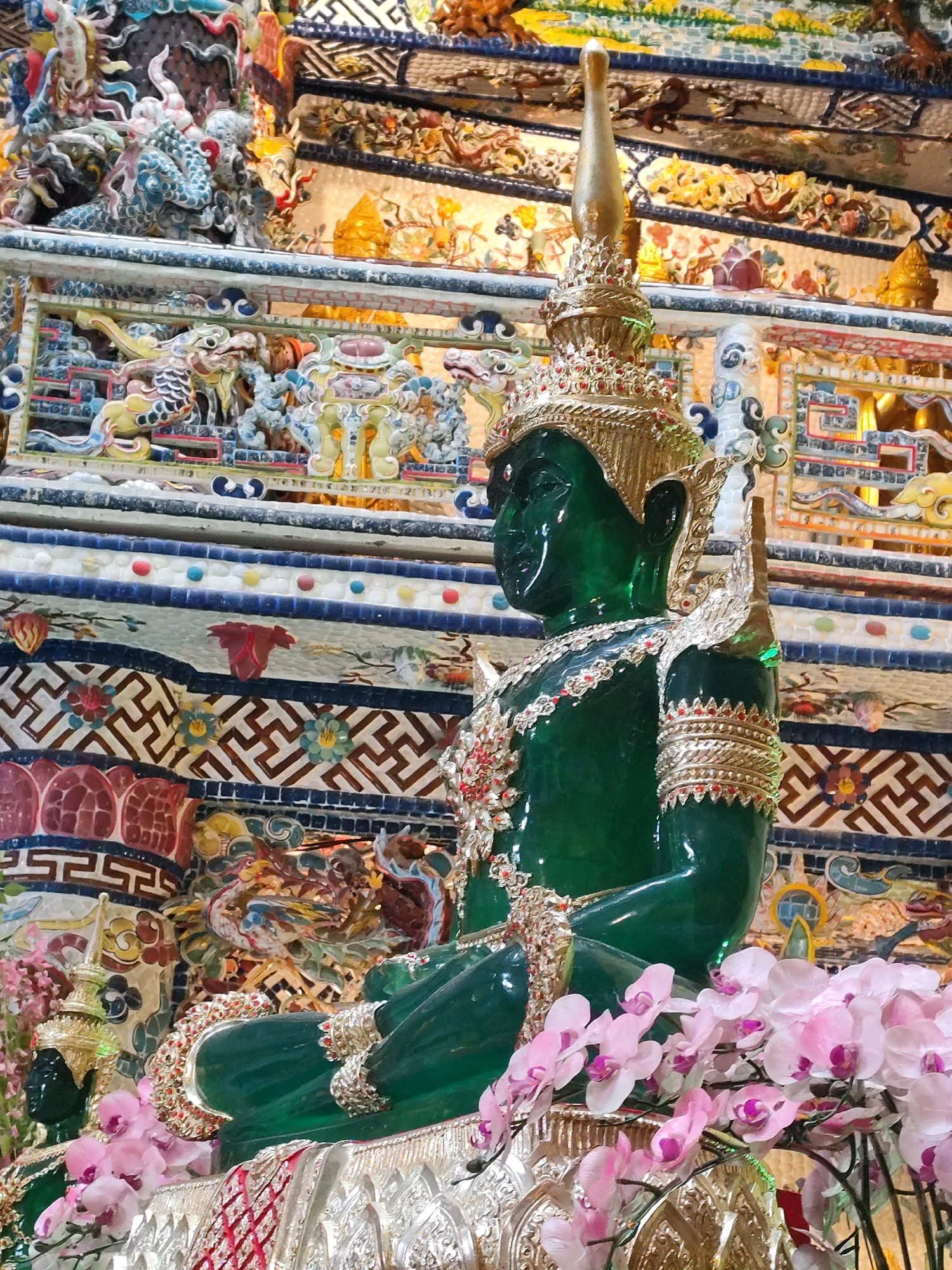 This image shows a close-up, angled view of a large, seated green Buddha statue adorned with gold and jewel-toned decorations. The statue is positioned in front of an elaborately decorated wall or altar, featuring intricate patterns and figures, likely in a temple setting within Xuân Thành, Vietnam. The statue itself is a vibrant emerald green, with a golden headdress and ornate jewelry around its neck and arms. It is seated in a meditative pose, with its legs crossed. In the foreground, a cluster of delicate pink orchids spills over the base of the statue. The background is a tapestry of colorful, mosaic-like decorations that include depictions of mythical creatures and geometric patterns. The overall impression is one of reverence and opulence.