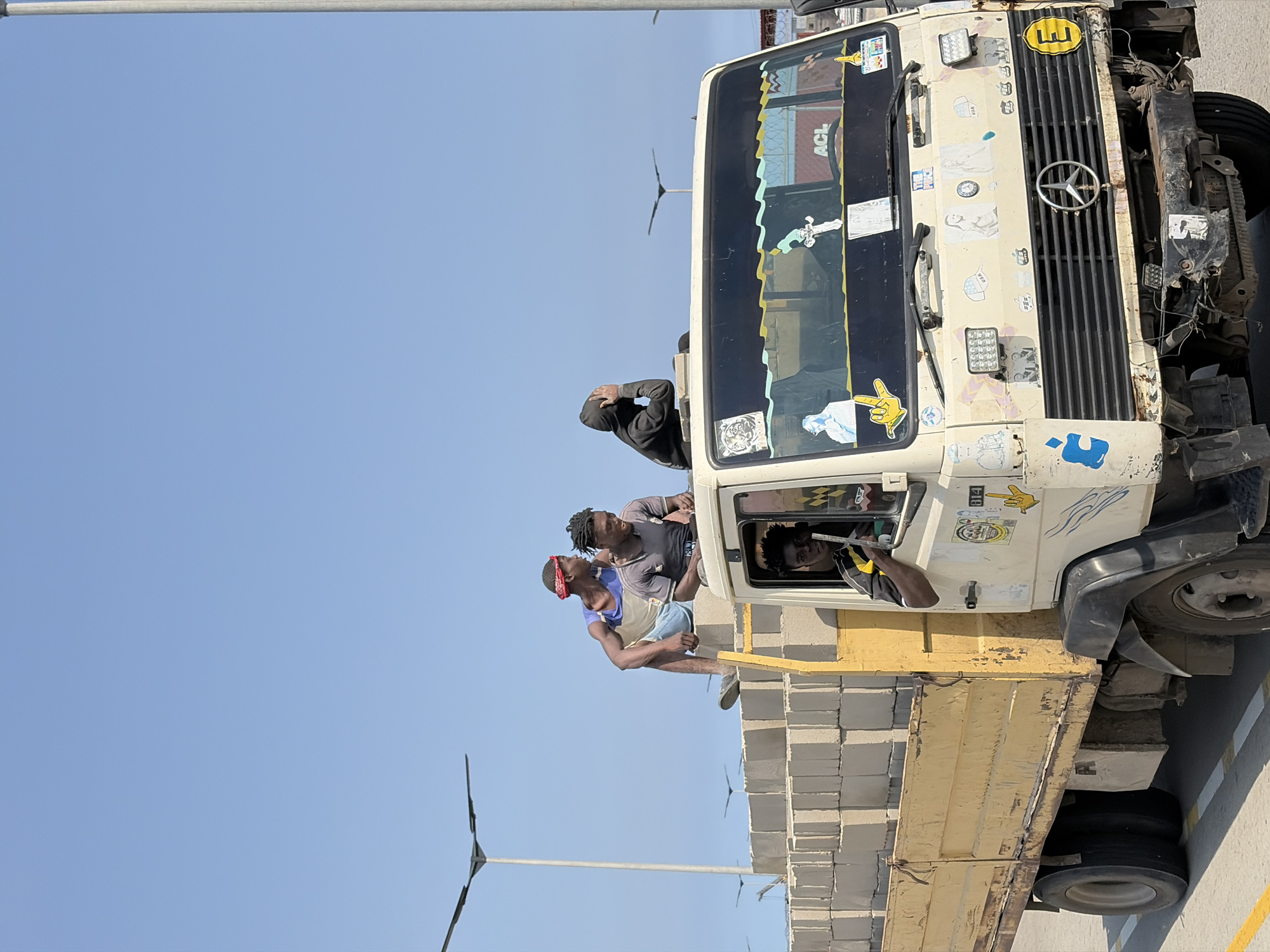 The image depicts a white Mercedes-Benz truck carrying a load of cinder blocks. Several people are present on or near the truck. Two individuals are sitting on top of the cinder block load, while another person is visible in the driver's seat. A person wearing a dark hoodie leans out from the front of the truck. The truck is parked on pavement. Wind turbines in the distance hint at Alaguntan, Nigeria.