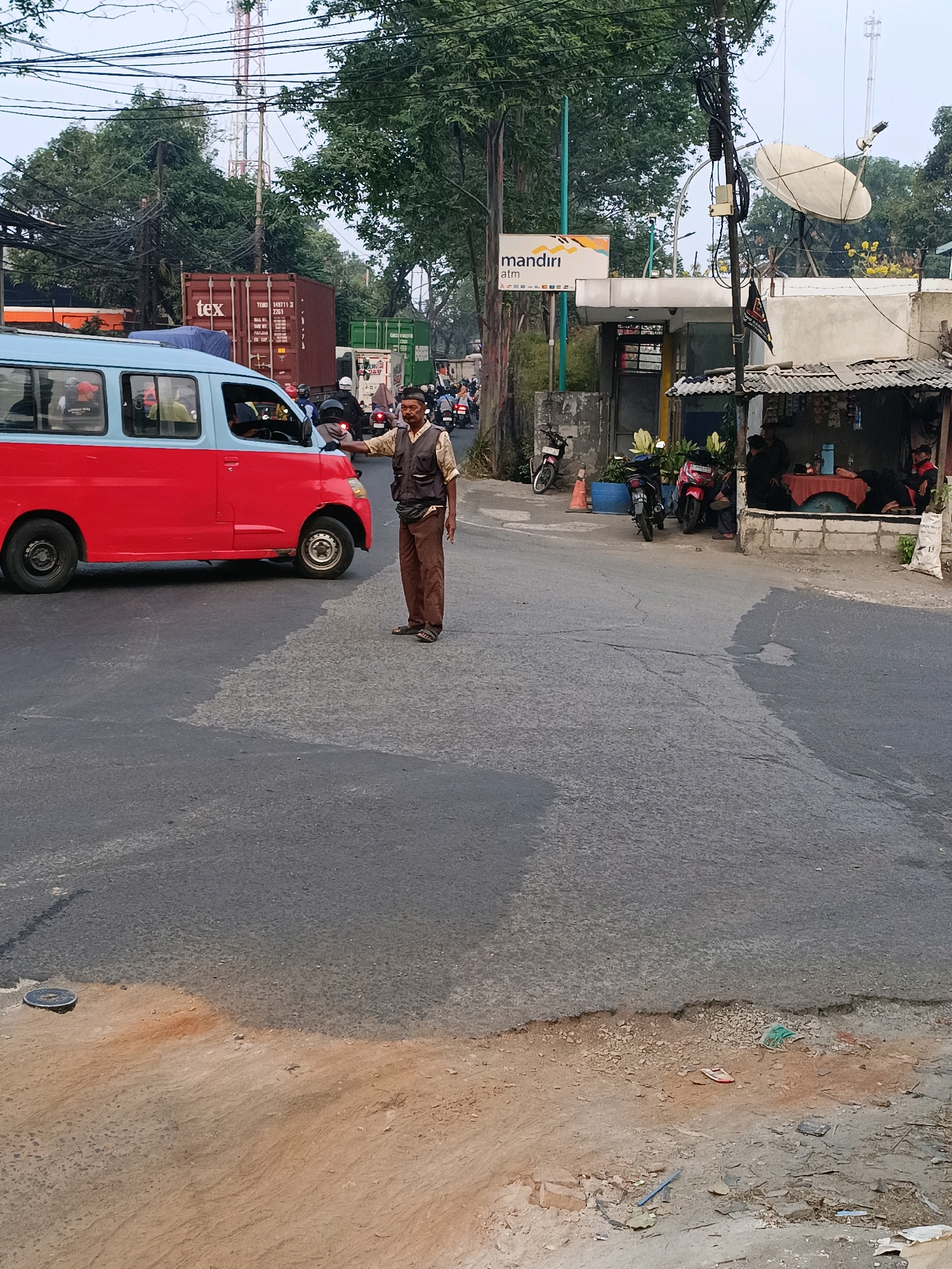 The image depicts a street scene in Tangerang, Indonesia, characterized by a busy road with traffic, including a red and blue van and various motorcycles. A man, possibly a traffic director or bystander, stands in the road, gesturing towards the van with his right hand. He is wearing a dark vest over a light-colored shirt and brown pants. Several motorcycles are parked or moving in the background. To the right, a small structure with a corrugated metal roof appears to be a roadside stall or kiosk, with people seated inside. A satellite dish is visible on a building behind the stall. The environment shows signs of urban development with visible power lines and trees lining the street. The road surface shows wear and tear, with patches of different textures and some debris on the roadside. The overall impression is one of a lively, everyday urban environment in Indonesia.