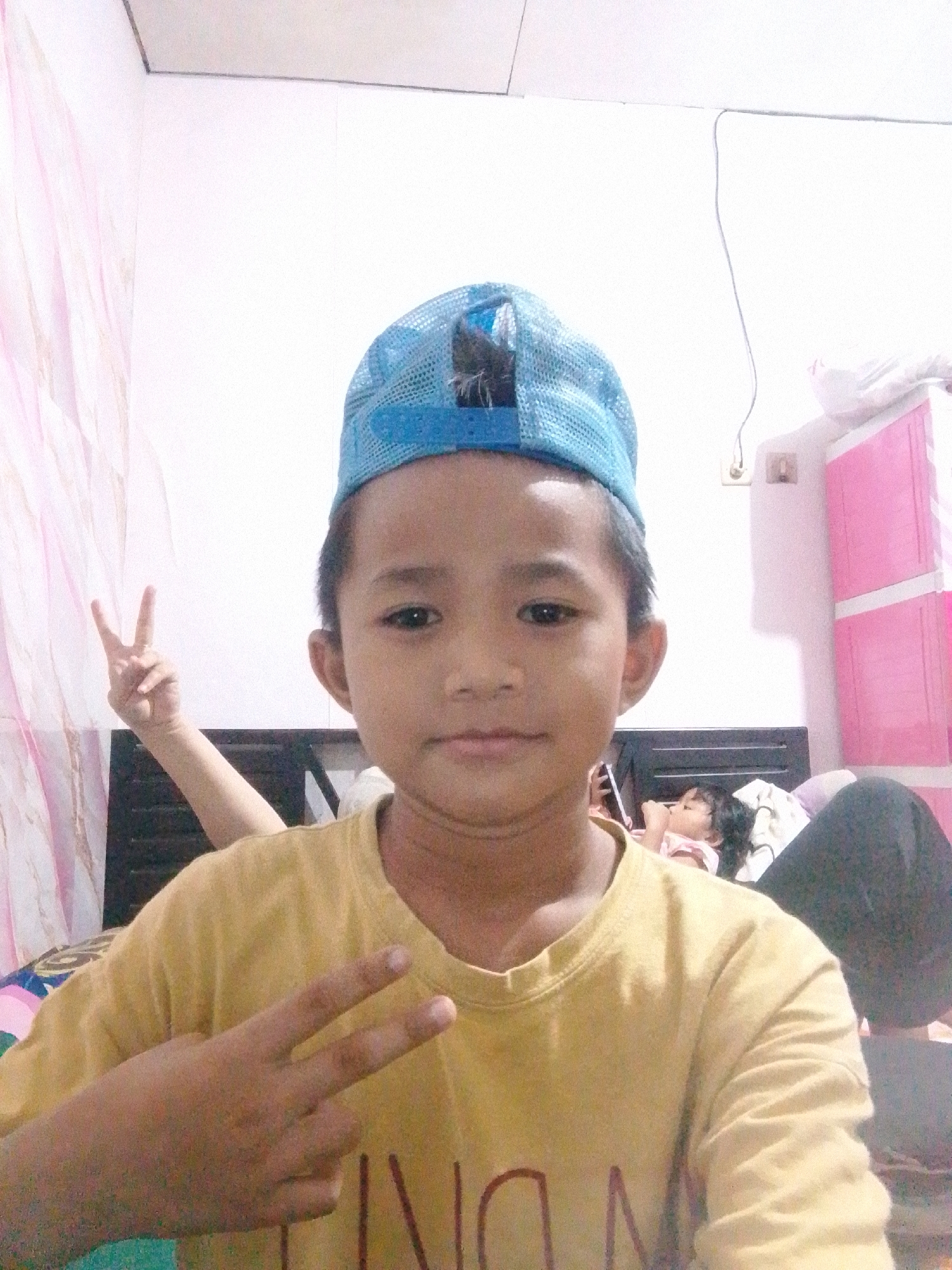 This is a frontal, waist-up, medium-shot selfie of a young boy wearing a blue mesh baseball cap and a yellow t-shirt. He is positioned in the center of the frame, facing the camera and making a peace sign with his left hand. His right arm is bent, with his hand also forming a peace sign behind him. In the background, a young child is lying down on a bed, with another person's legs visible on the right side of the frame. A pink, multi-tiered cabinet is visible on the far right. The setting appears to be a bedroom, likely in a residential area of South Jakarta, Indonesia, given the style of the cabinet and the general ambiance.