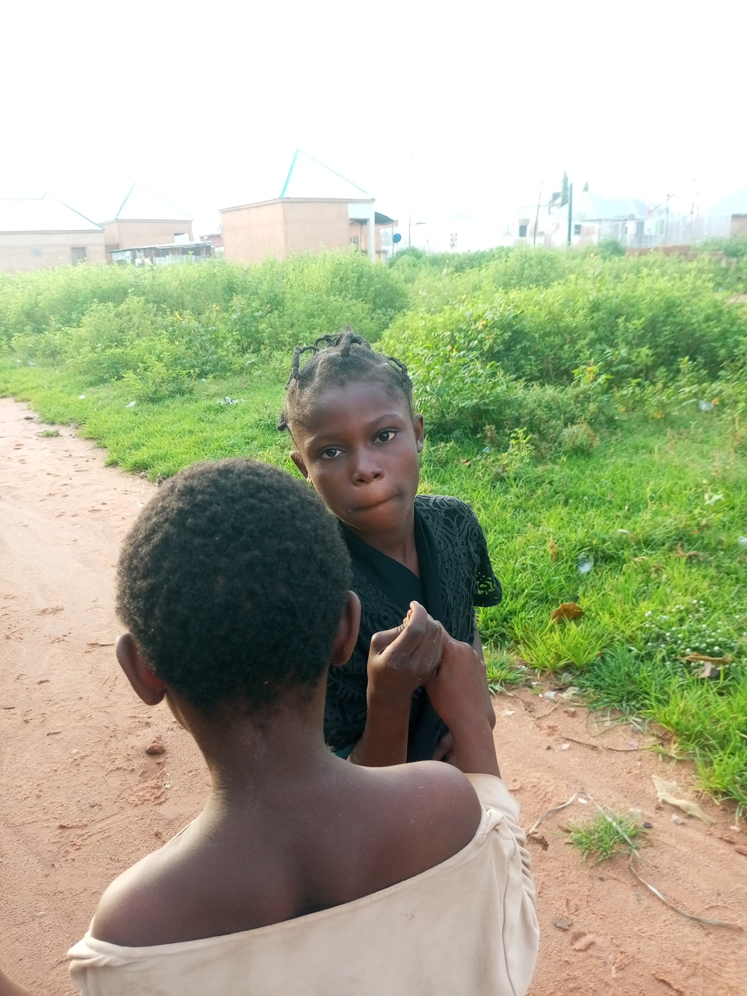 Two young girls are the subjects in this outdoor photo. One girl, with her back to the camera, has short, dark, curly hair and is wearing a light-colored off-the-shoulder top. Her skin appears to have some dryness or flakiness on her neck and shoulders. The other girl is facing the camera, looking directly at the viewer with a serious expression. She has dark skin and her hair is styled in braids. She is wearing a black, possibly lace, top. Her hands are clasped together in front of her.
The setting appears to be a rural or semi-rural area in Tempure, Nigeria. The ground is sandy and a dirt path is visible. Lush green vegetation and tall grasses are abundant in the background, with some scattered debris. In the far distance, there are hints of buildings with light-colored walls and light blue or green roofs, suggesting a developed area nearby. The sky is overcast, and the lighting suggests it is daytime, possibly late afternoon or early morning, as there are no strong shadows. The overall mood is somewhat subdued, with the direct gaze of the girl facing the camera being a prominent feature. There is no visible text in the image.